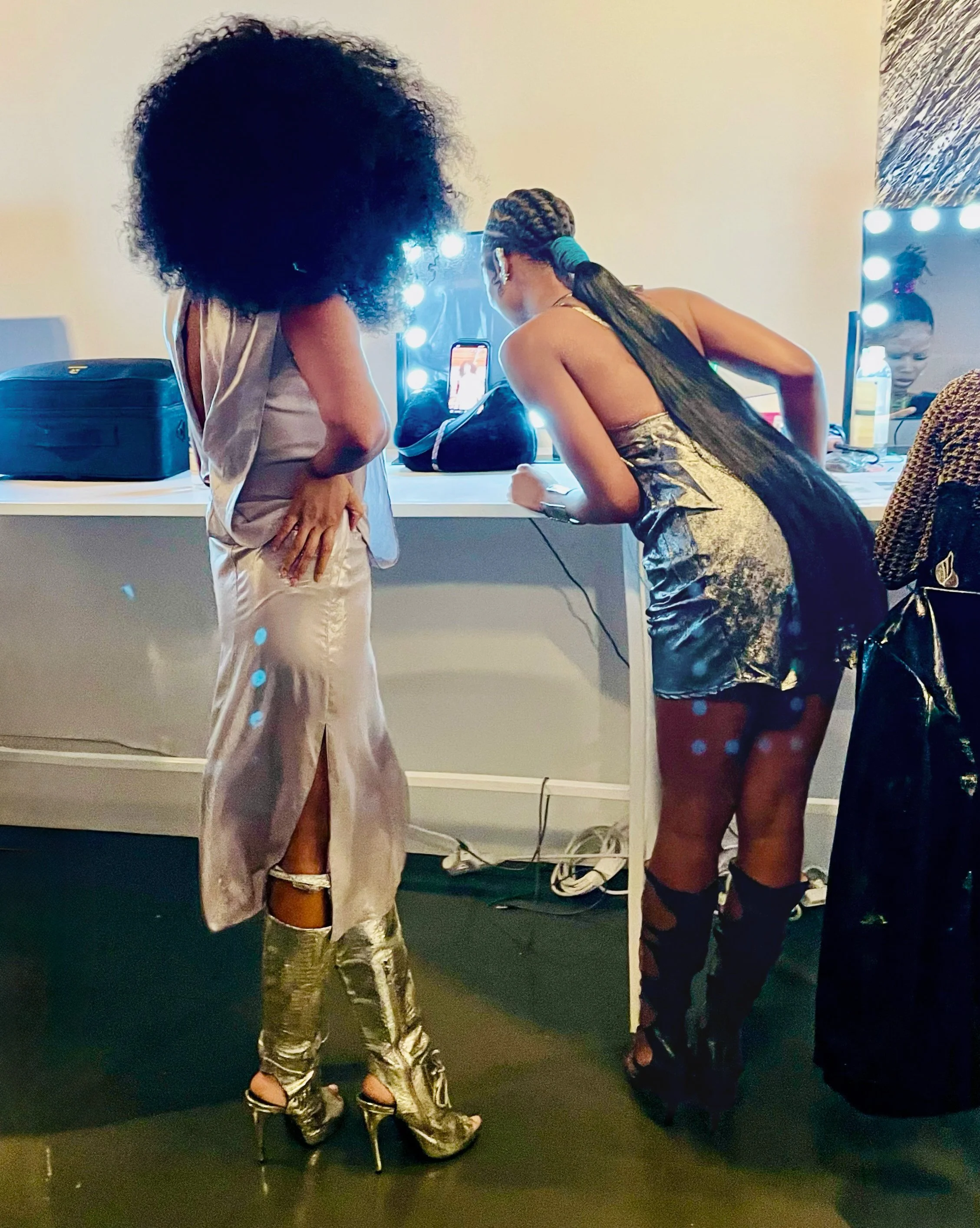Two women looking at a phone screen in front of a mirror with bright lights, one wearing a satin dress with high-heeled boots, the other in a shiny dress and leg warmers.