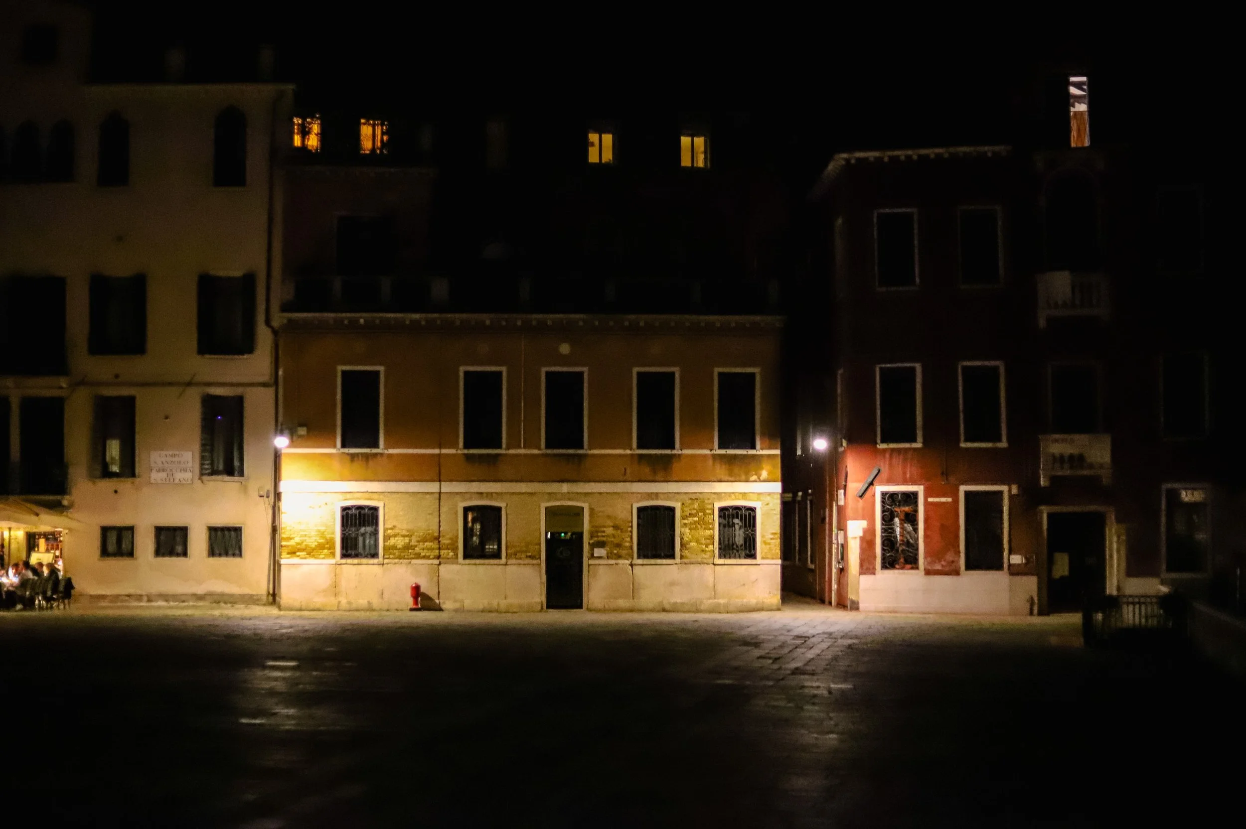 Nighttime view of a row of buildings with lit windows, with a small outdoor seating area on the left and a fire hydrant in front of the center building.