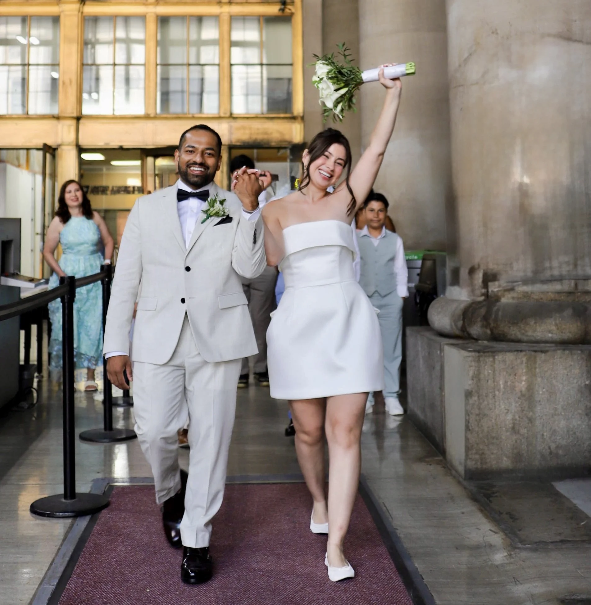 A joyful bride and groom walking hand in hand inside a building, with the bride raising a bouquet in the air and the groom smiling, surrounded by other wedding guests.