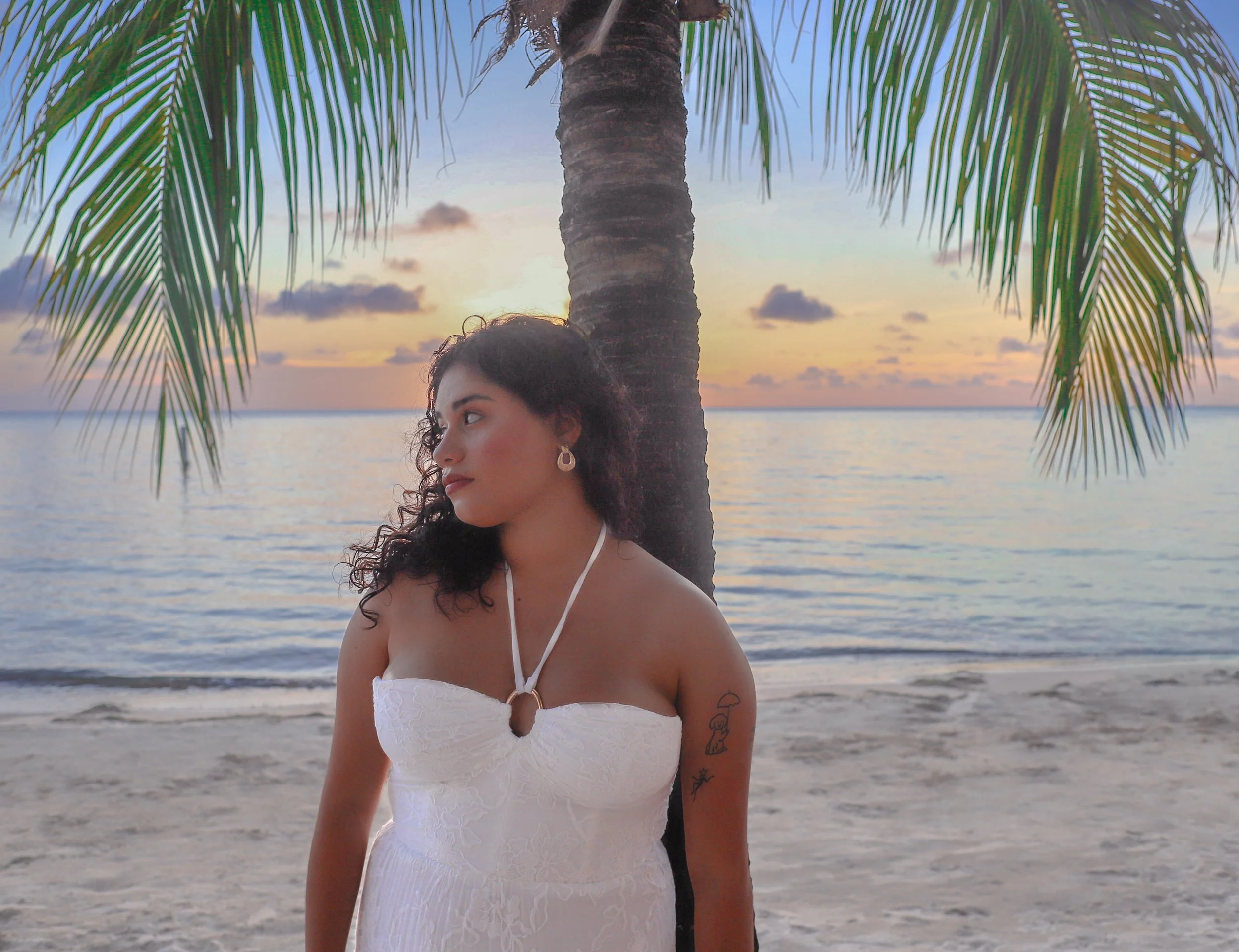 A woman with dark curly hair and hoop earrings stands on a beach next to a palm tree, looking to the left, during sunset with a colorful sky and calm ocean in the background.