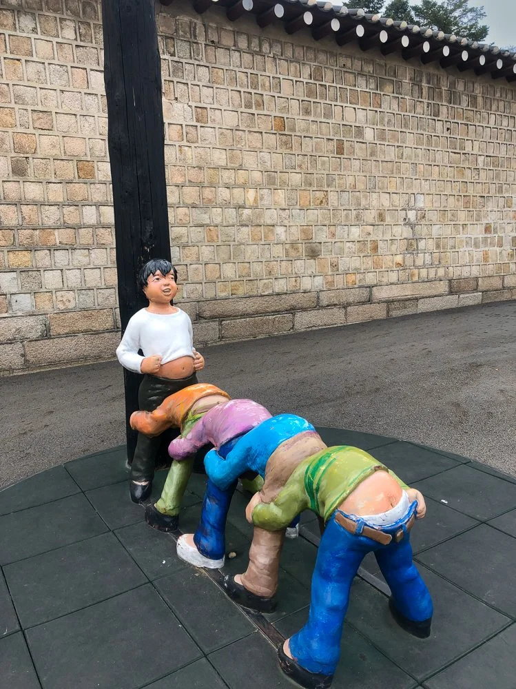 Statue of a laughing boy standing behind four colorful, clay-like statues of people bowing forward. The boy has black hair, wearing a white shirt and black pants. The statues are in various bright colors, mounted on a black tiled surface against a br
