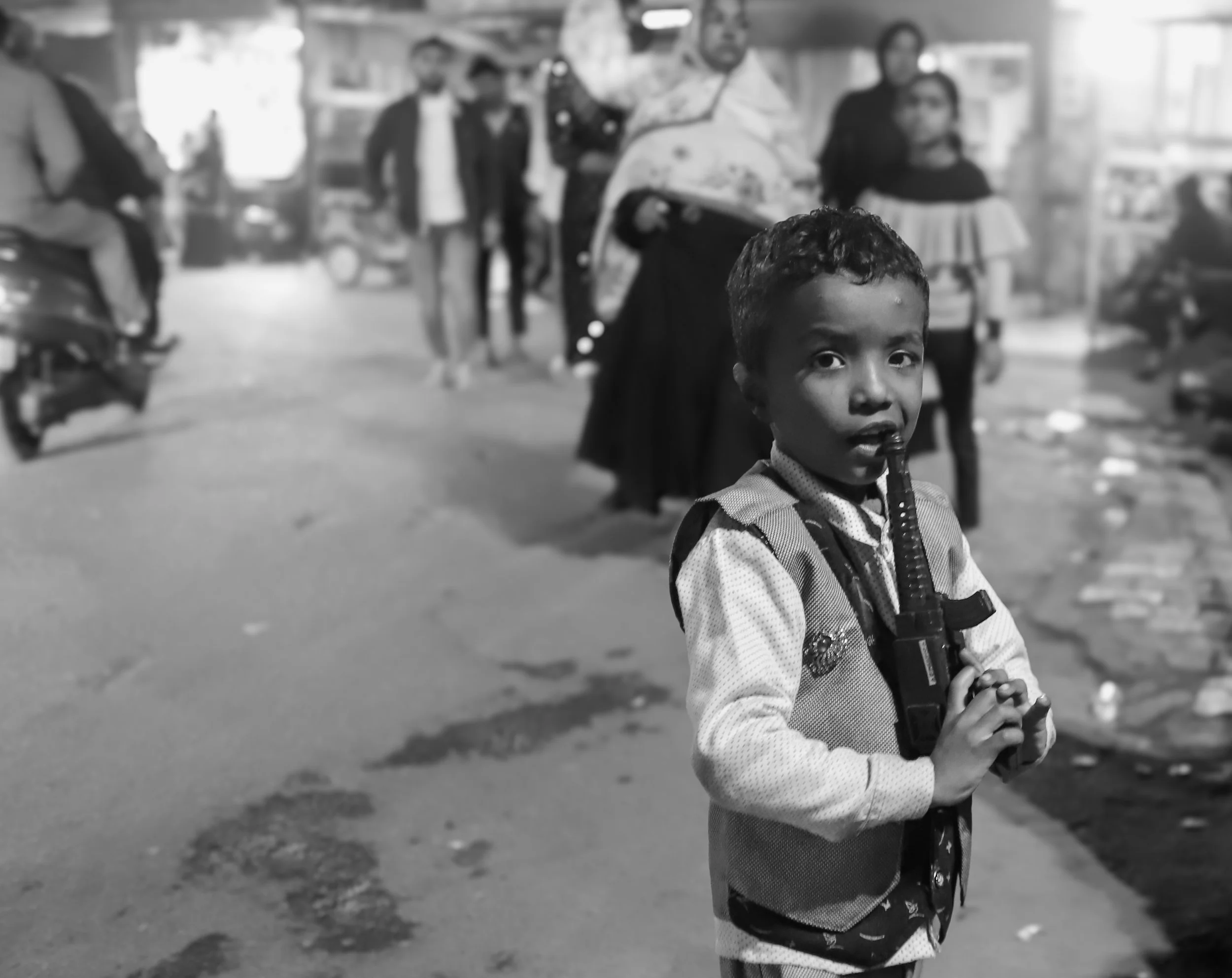 A young boy holding a toy gun on a busy street at night, with several people and vehicles in the background.