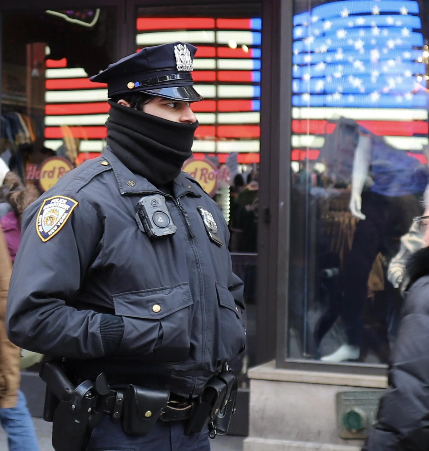 A police officer in uniform and a black face covering standing with hands in pockets outdoors, with American flag-themed window in background and pedestrians nearby.