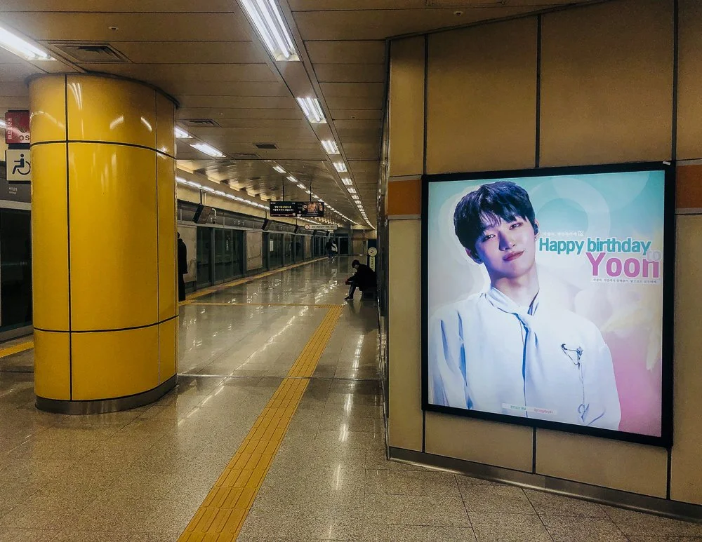 Indoor subway station platform with a yellow pillar on the left and a digital billboard on the right displaying a birthday message for Yoon featuring a young man with dark hair in a white shirt.