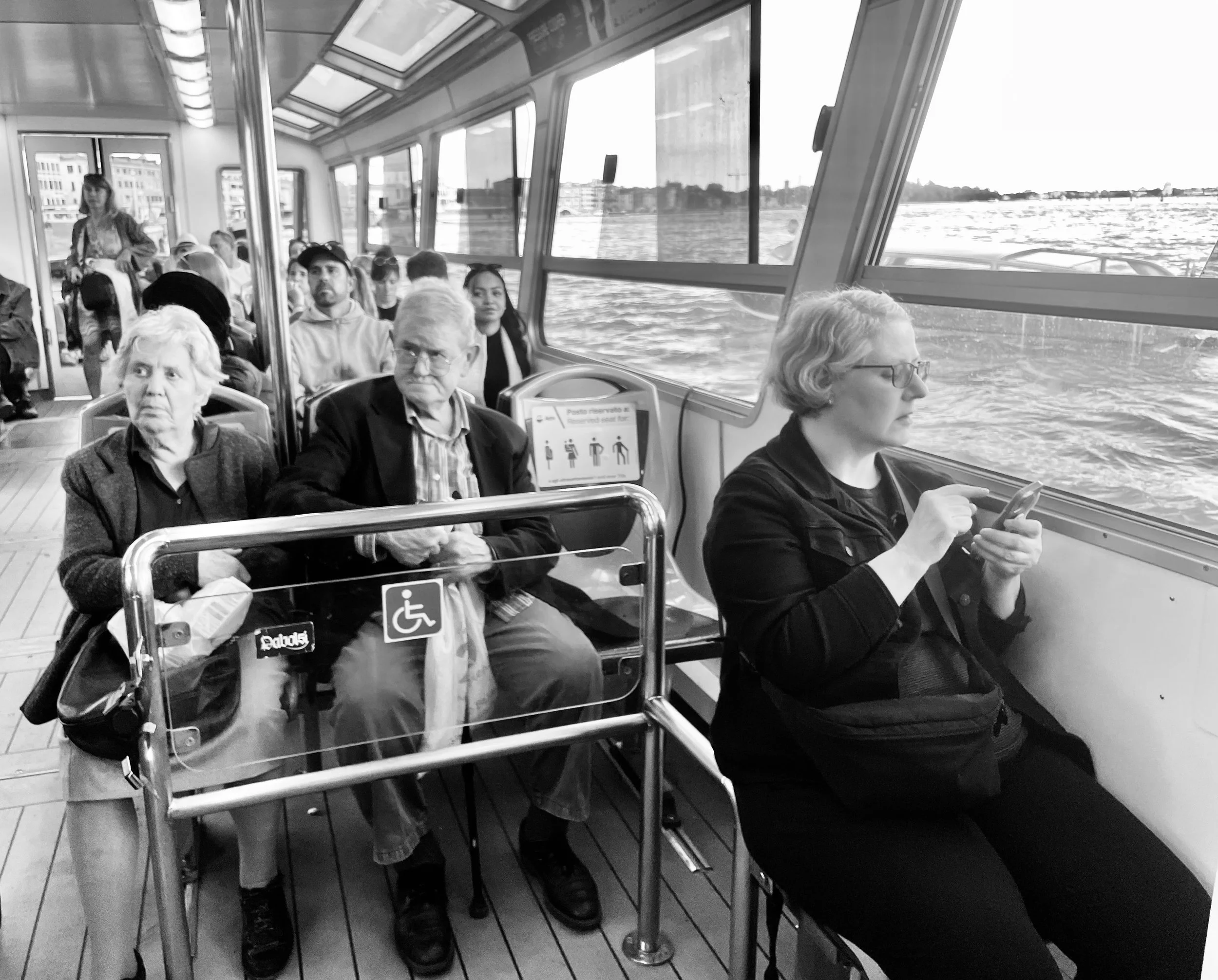 People seated inside a passenger boat or ferry, with water and buildings visible through large windows. Some passengers are looking out or using their phones, including an older woman and man in the foreground, and a woman in the background.