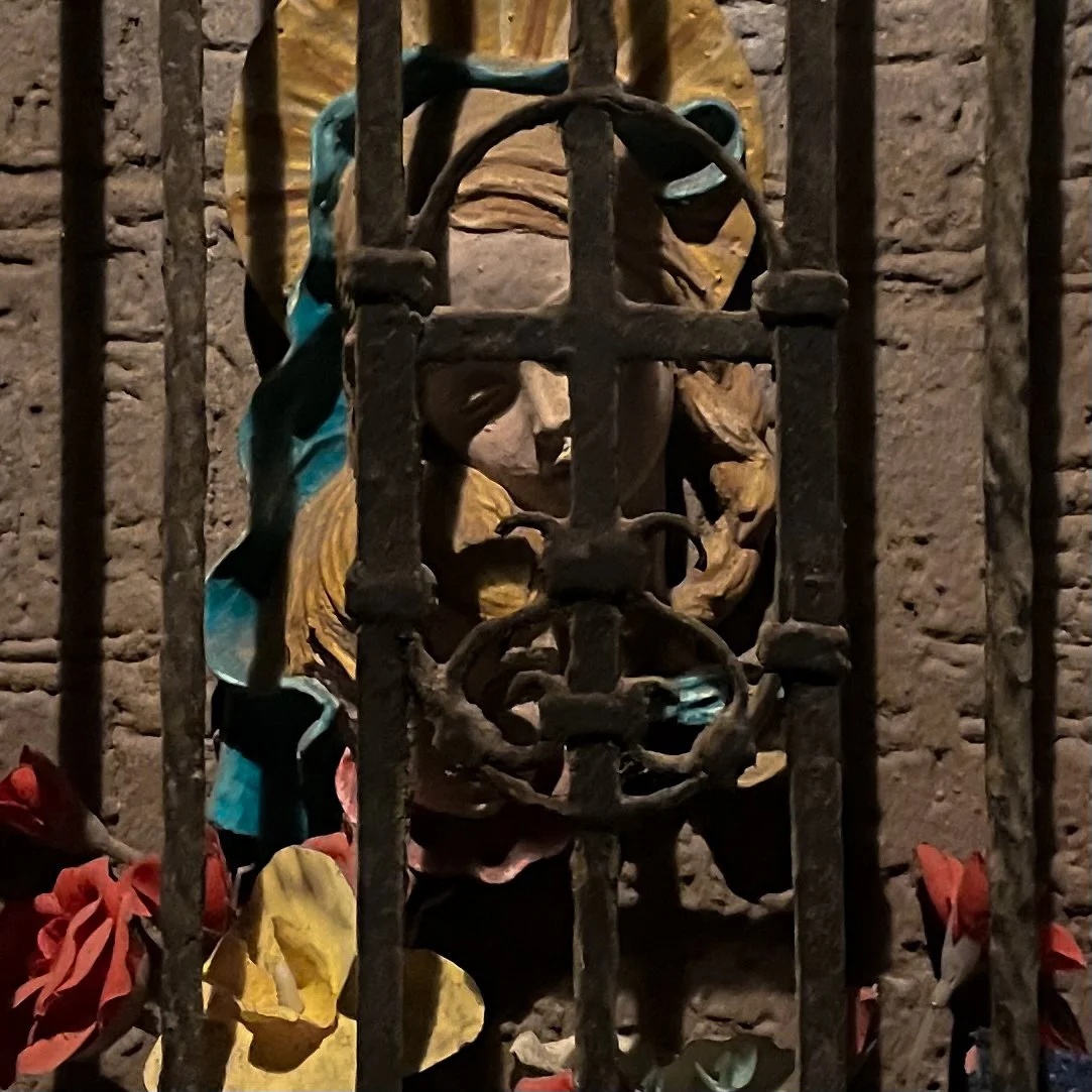 A religious statue behind metal bars, featuring a figure with a golden crown, surrounded by colorful flowers and greenery.