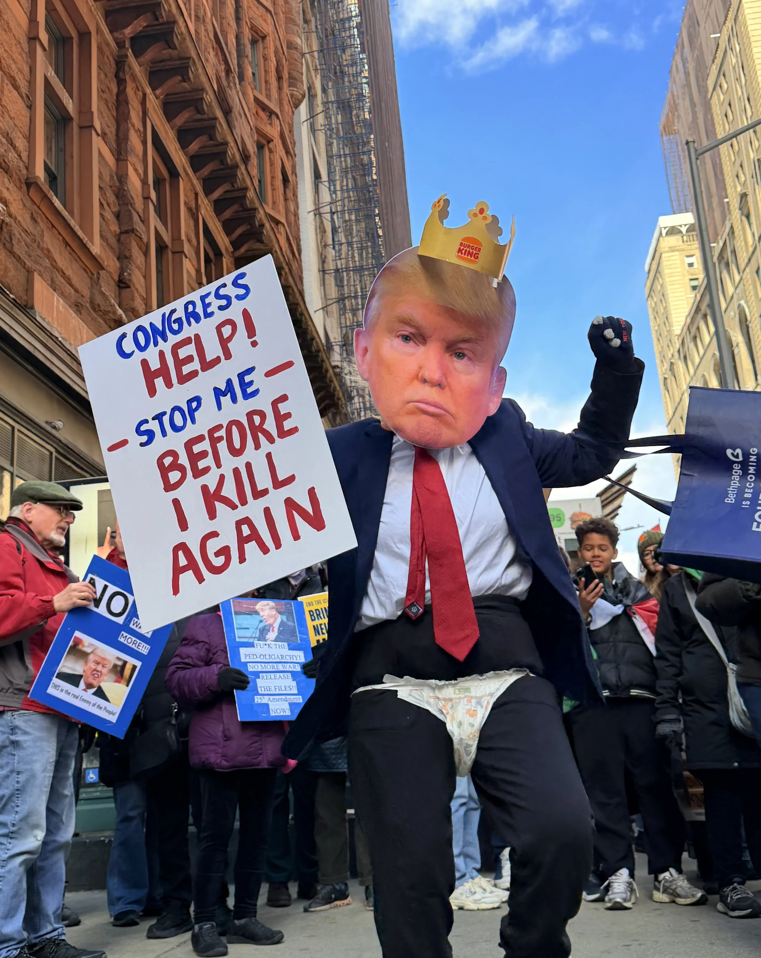 A person dressed in a suit and tie with a mask resembling Donald Trump, wearing a Burger King paper crown, holding a protest sign that reads "Congress Help! Stop me - before I kill again". The person is in a crowd of protesters in an urban area.