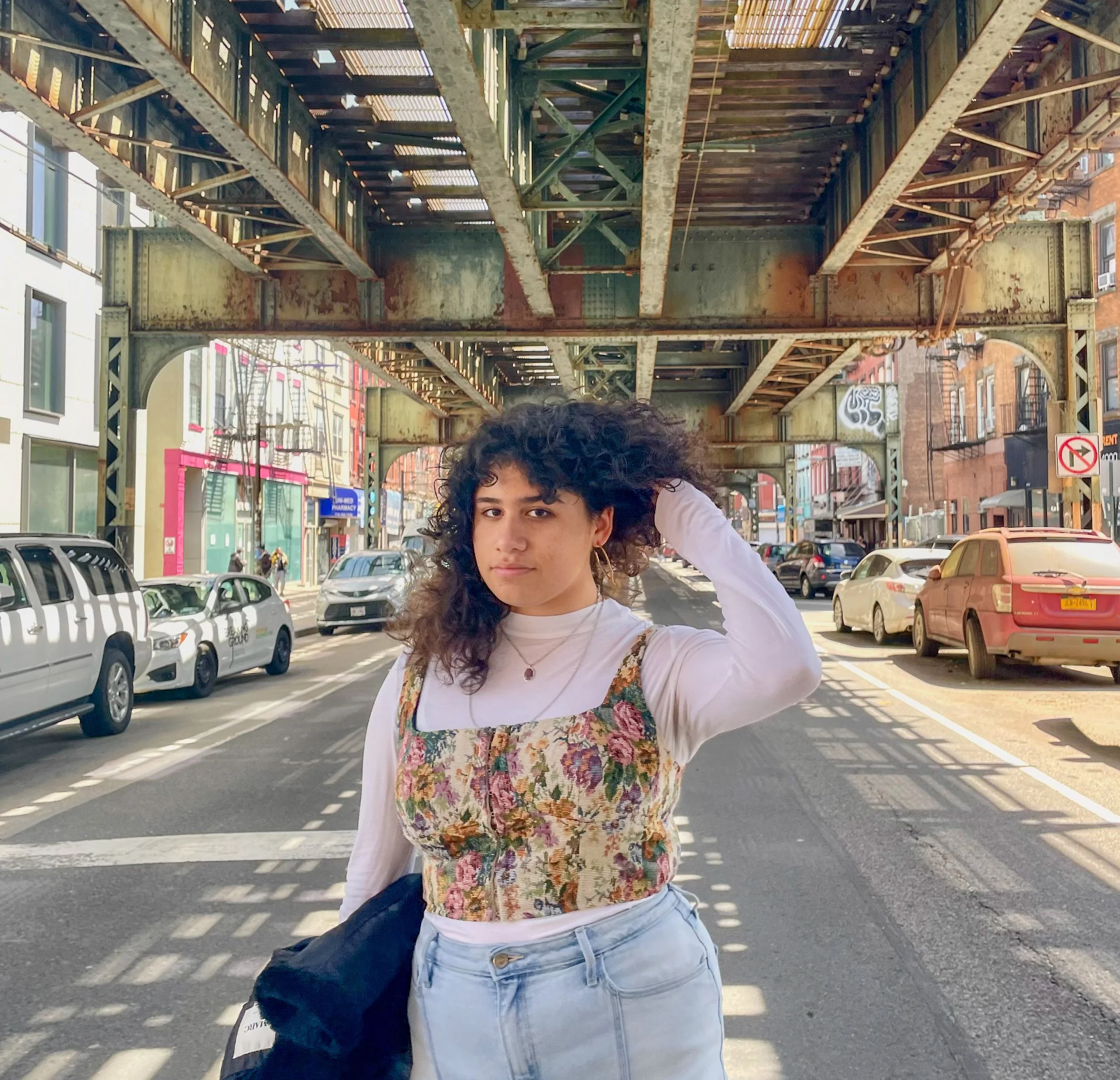Young woman with curly dark hair standing on city street under an elevated train track, wearing a floral top layered over a white long-sleeve shirt and light blue jeans.