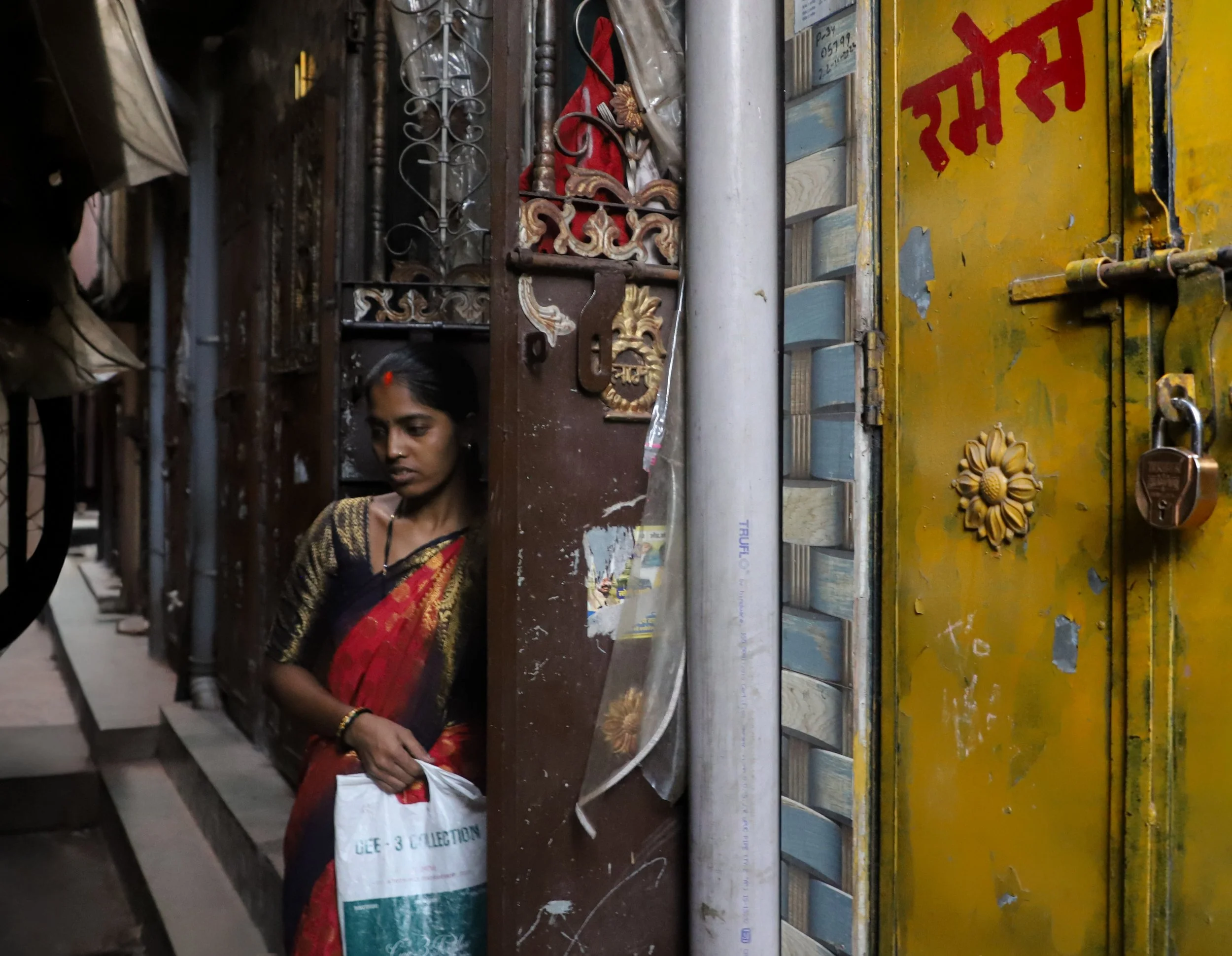 A woman in traditional Indian attire standing near a partially open gate, holding a white bag, with a yellow painted metal door with a lock on it in an alleyway.