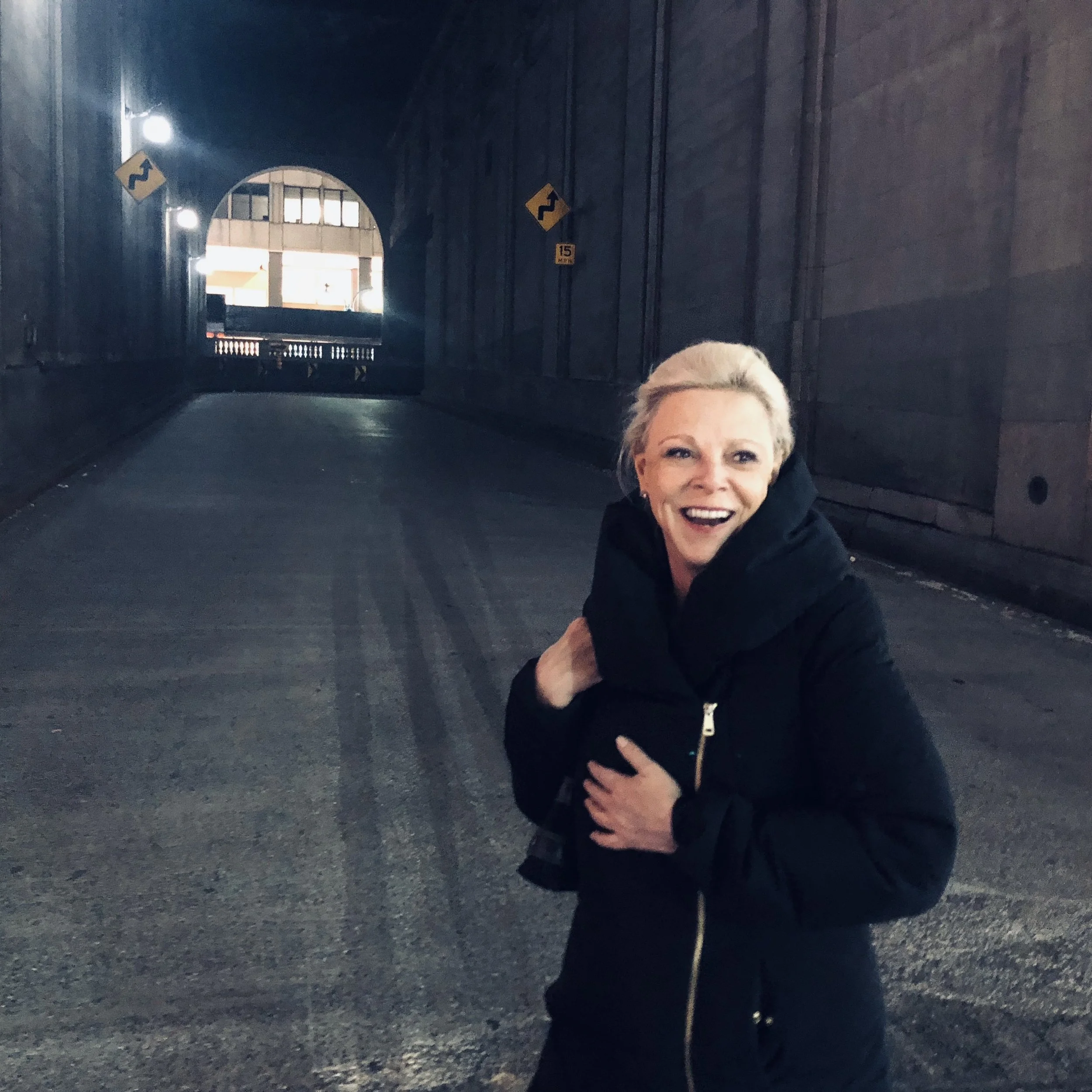 A woman with short, light-colored hair smiling, wearing a black jacket, standing in a dark underpass or tunnel with lights on the side, and a bright opening in the background.