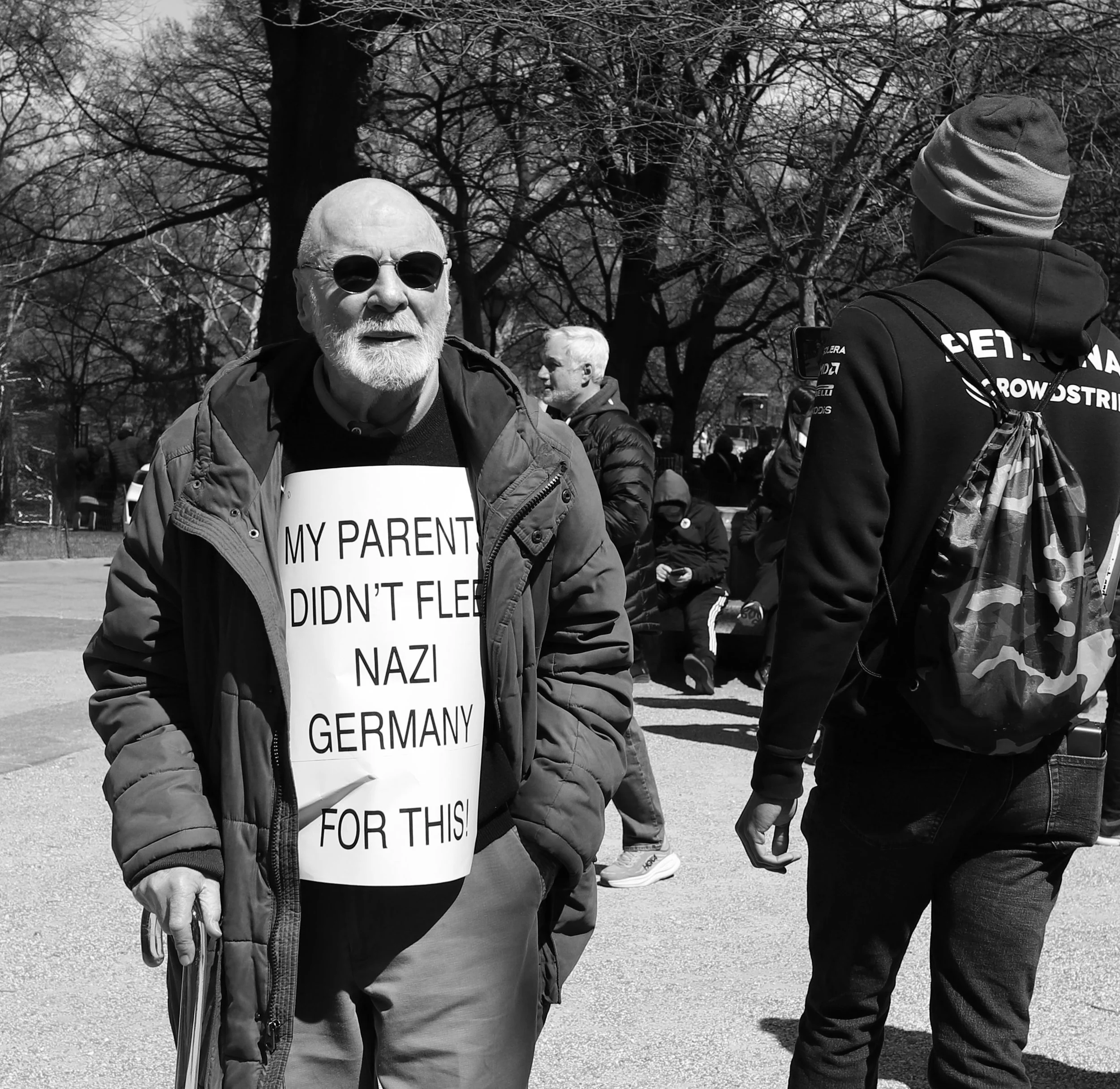 An elderly man wearing sunglasses and a puffy coat has a sign on his chest that reads, "My parent didn't flee Nazi Germany for this!" He is standing outdoors in a park with leafless trees and other people in the background.
