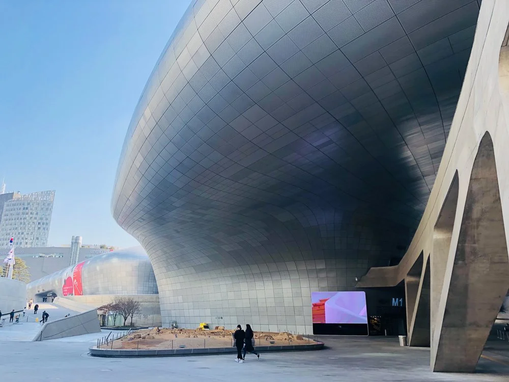 Modern architectural building with a curved metallic facade and an outdoor digital screen. A couple walks nearby in an urban setting with a clear blue sky.