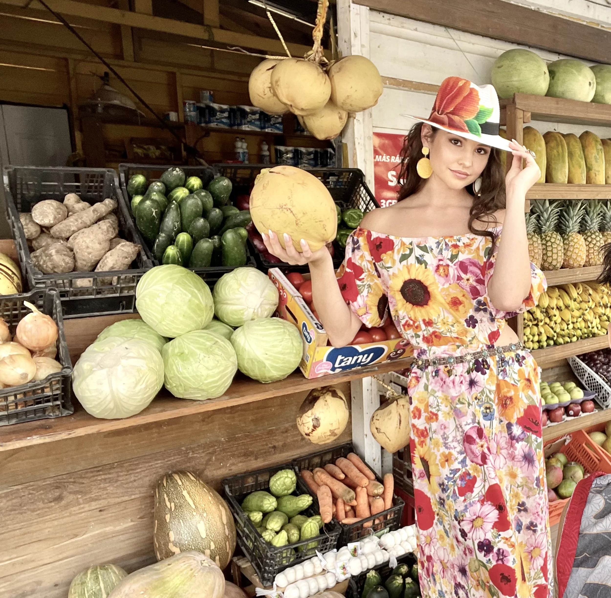 A woman in a colorful floral dress and matching hat stands in a market stall filled with various fresh vegetables and fruits, holding a yellow melon in one hand and adjusting her hat with the other. Roatan.