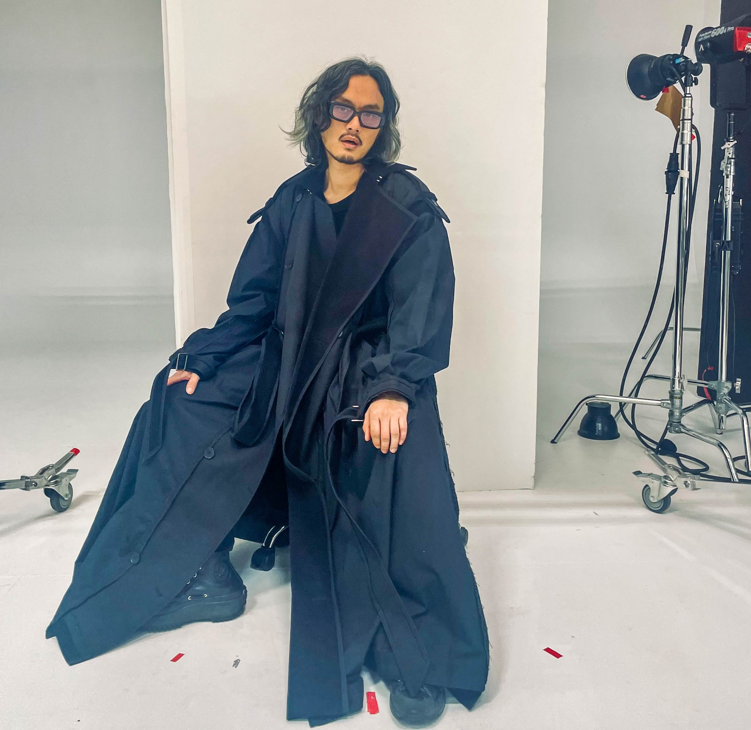 Person with long hair and glasses sitting on a stool in a photo studio, dressed in black with large black coat, surrounded by lighting equipment and a white backdrop.