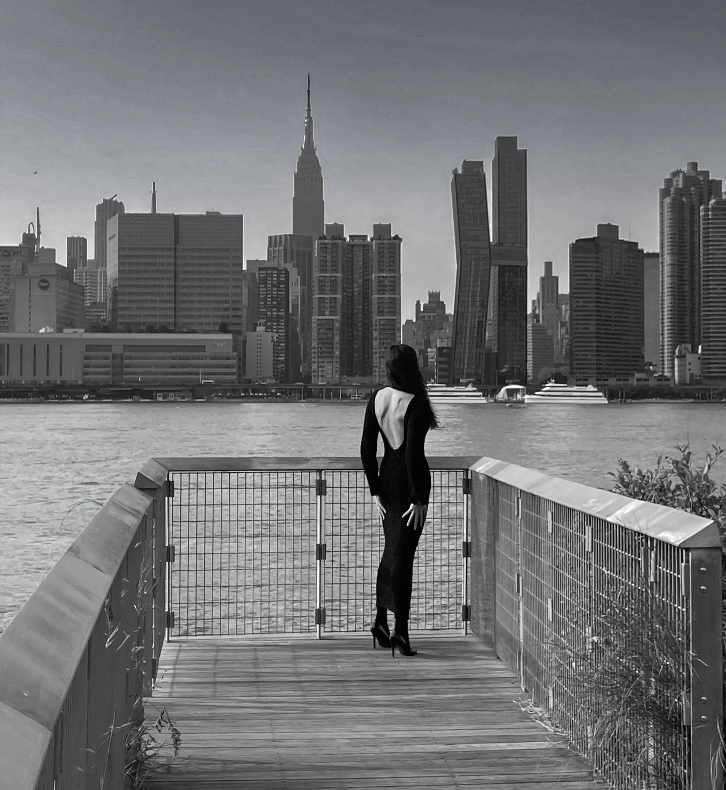 A woman in a long black dress and heels standing on a wooden walkway with a metal railing, overlooking a river with a city skyline, including the Empire State Building, in the background.