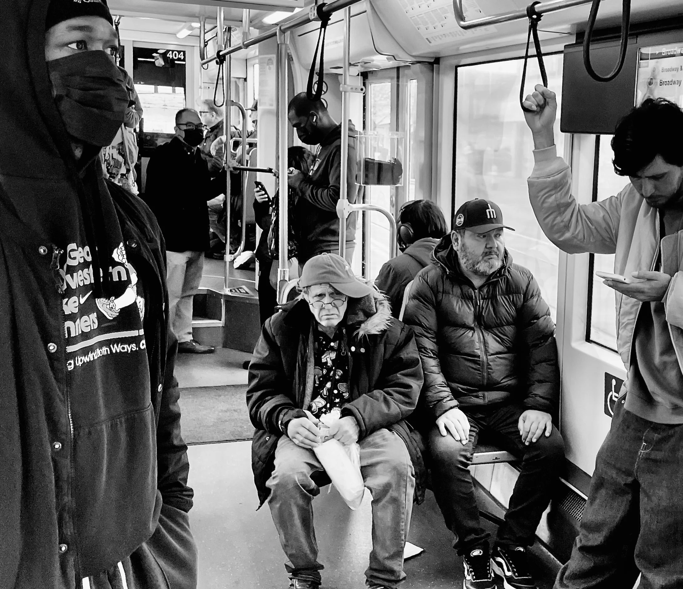 Passengers sitting and standing on a bus, wearing masks, with some using their phones, and a woman with glasses and a hat sitting on a seat looking forward.