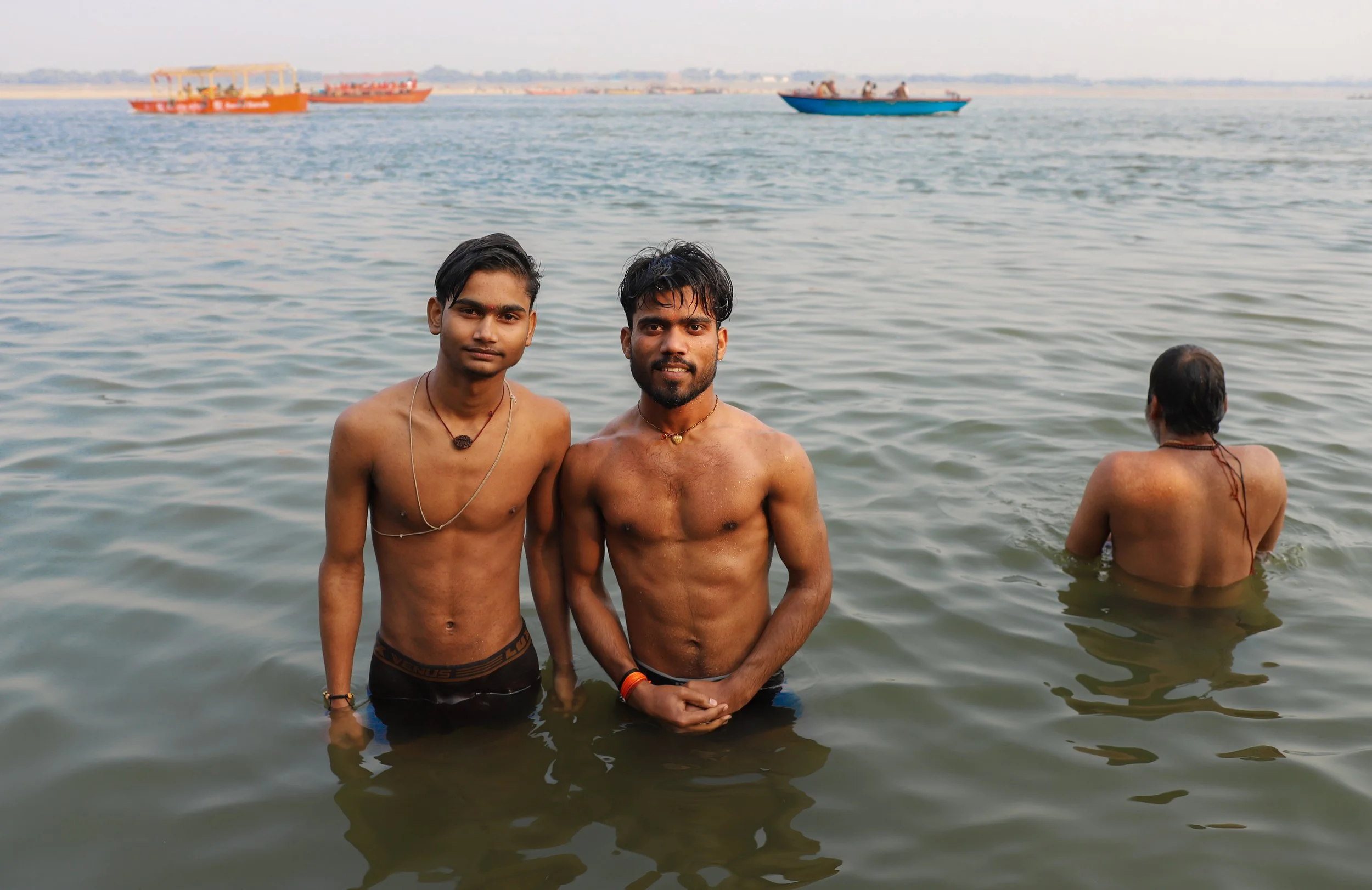 Two young men standing in a body of water, with one person swimming away from the camera in the background, and boats on the water, with a distant shoreline visible.