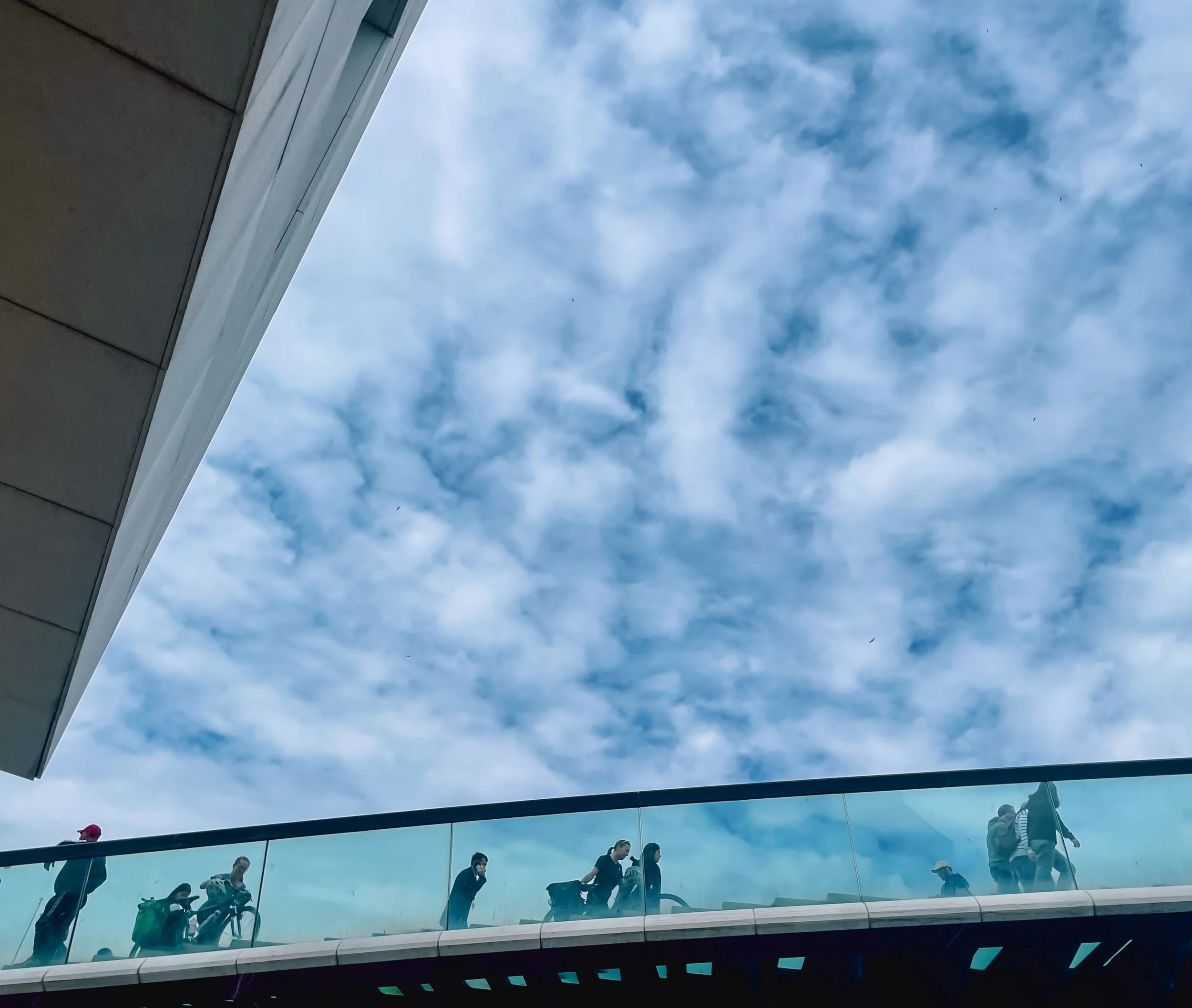 People walking on a glass bridge or walkway outside a building, with a blue sky and clouds above.
