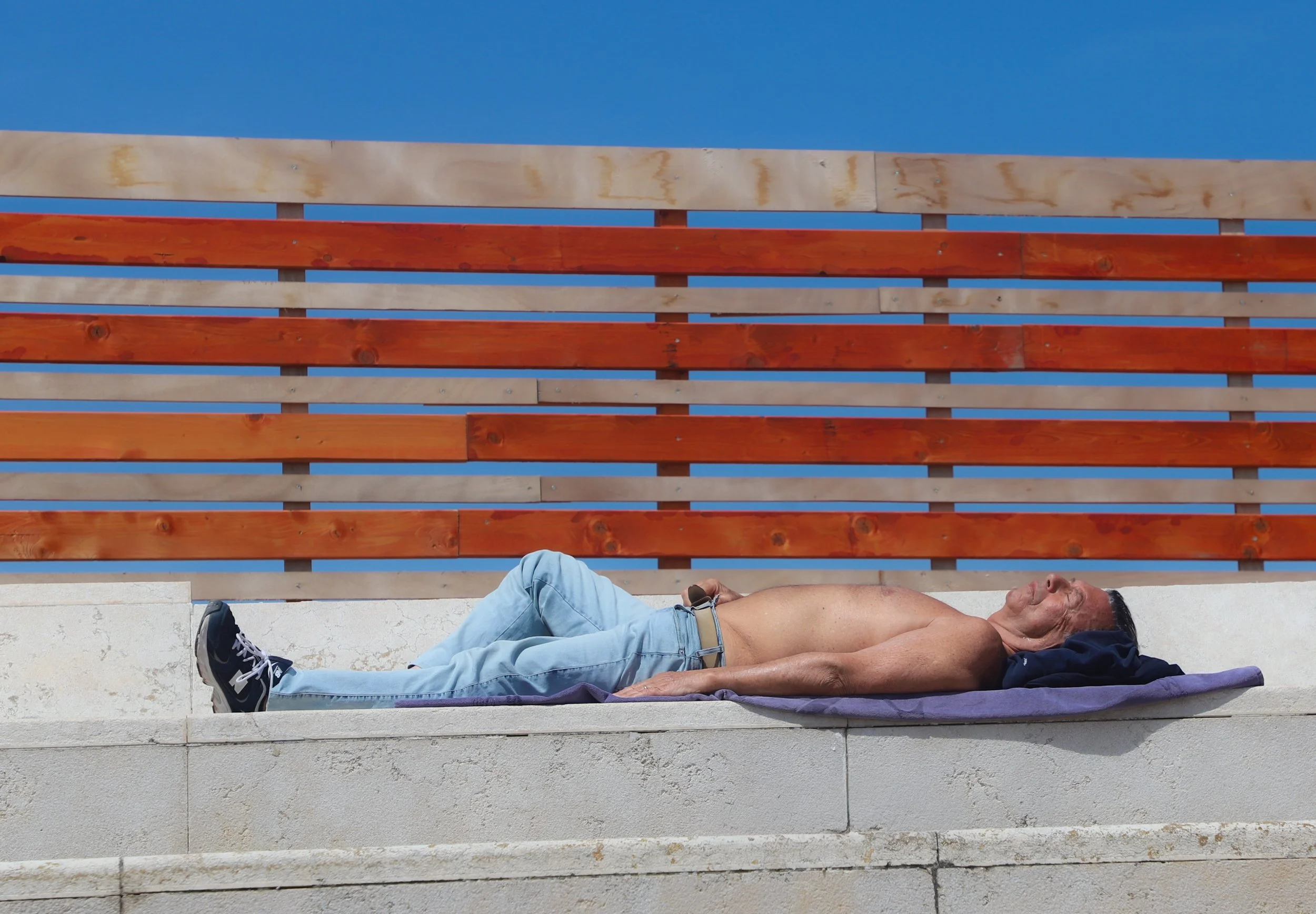 An older man lying shirtless on a purple towel on a concrete surface outdoors, wearing light blue jeans, black sneakers, with his eyes closed and resting under a clear blue sky, outlined by a wooden fence.