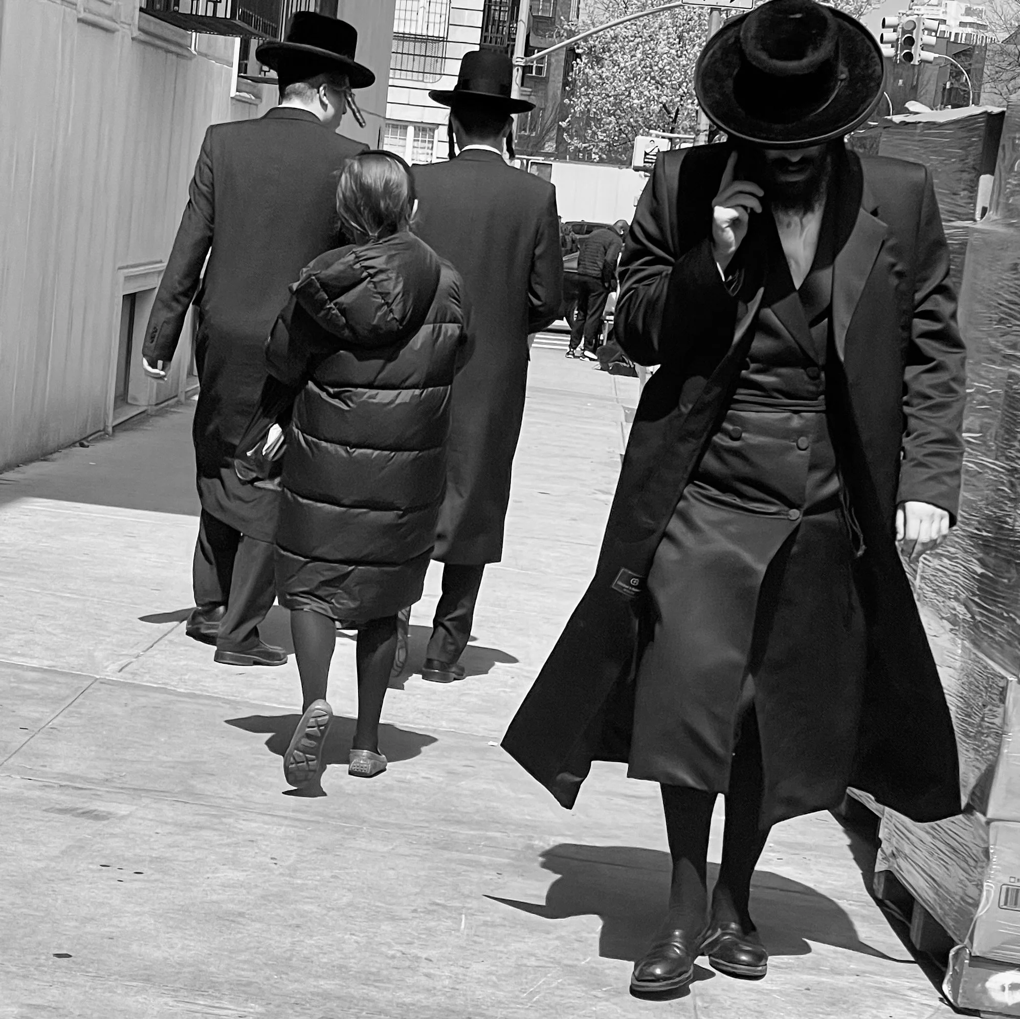 A black and white photo of four people walking on a city sidewalk. Three men are dressed in suits and wearing wide-brimmed hats. A woman is wearing a puffy coat and glasses.