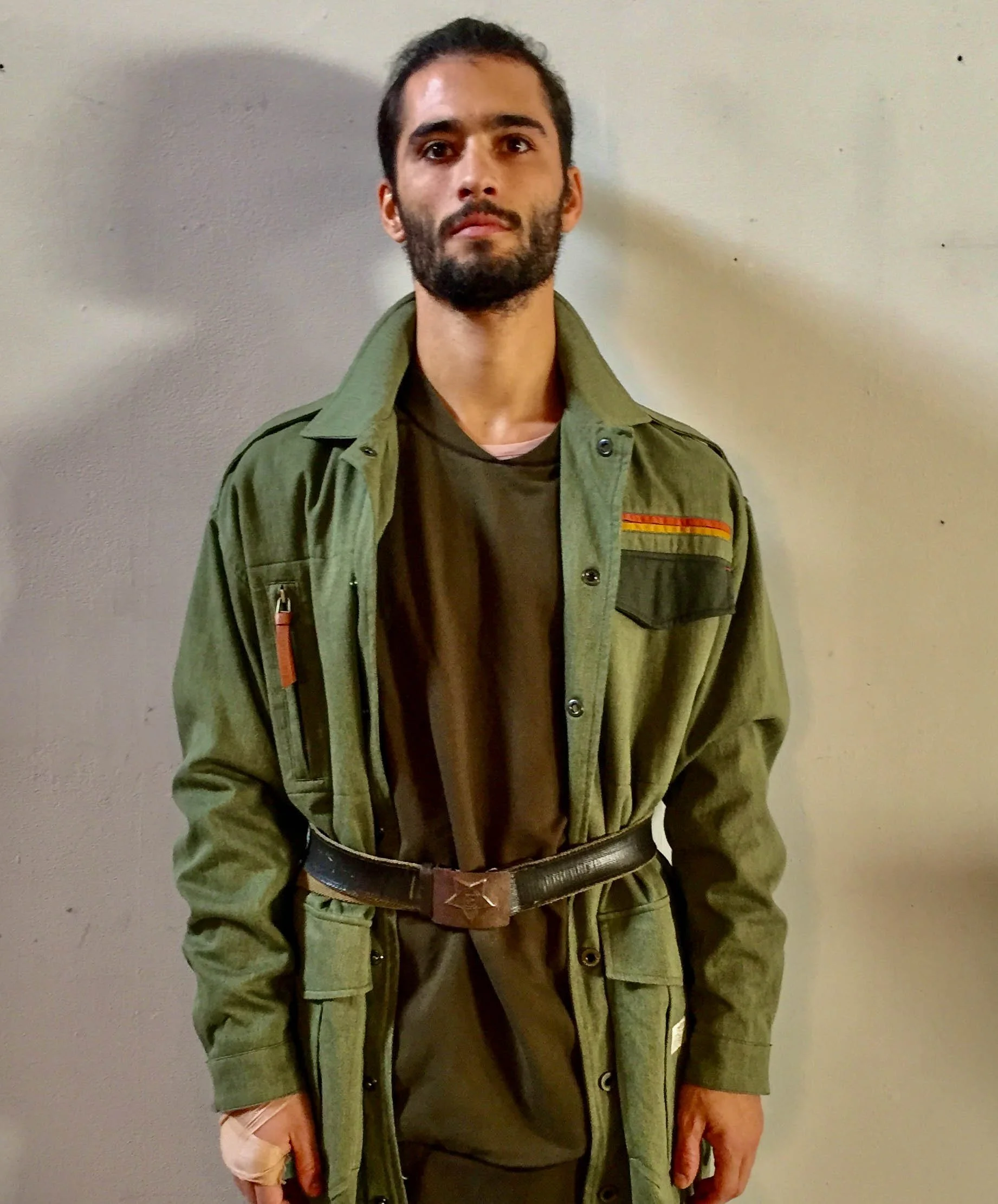 A man with dark hair and beard wearing a green military-style jacket with patches and a belt, standing against a plain wall.