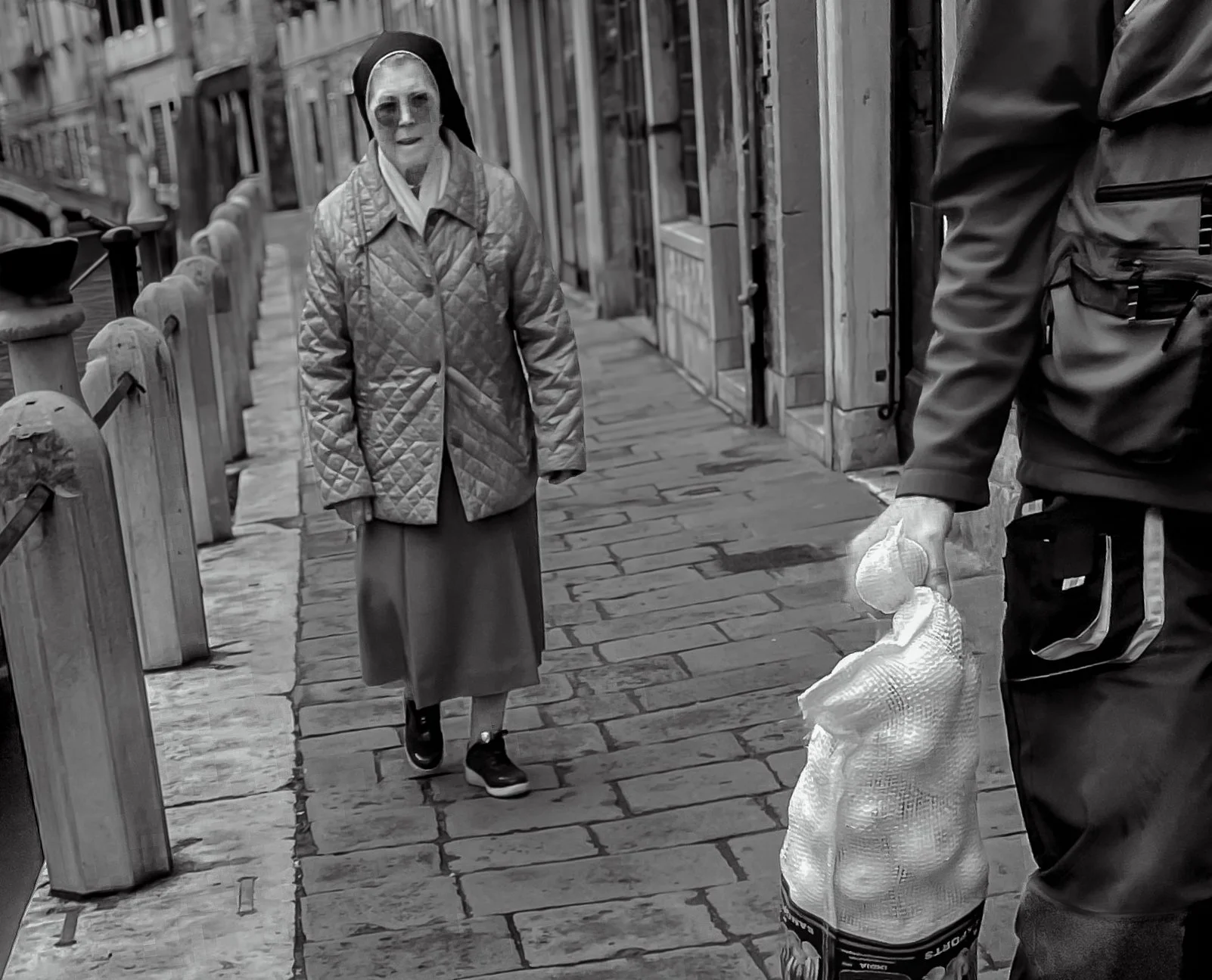 A woman wearing a quilted coat and a long skirt walks on a city sidewalk, while a man holding a bag of garlic reaches out to her. The scene is in black and white.