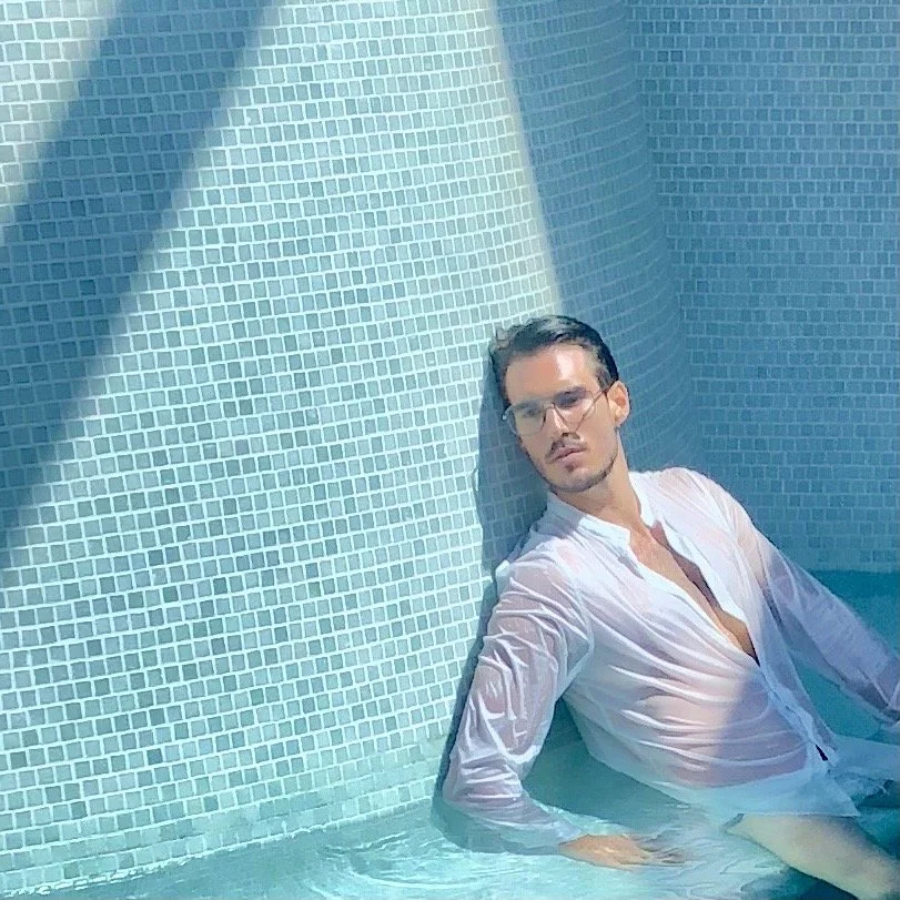 A man with glasses and wet hair, wearing a white shirt, sitting in a swimming pool with blue tiled walls.