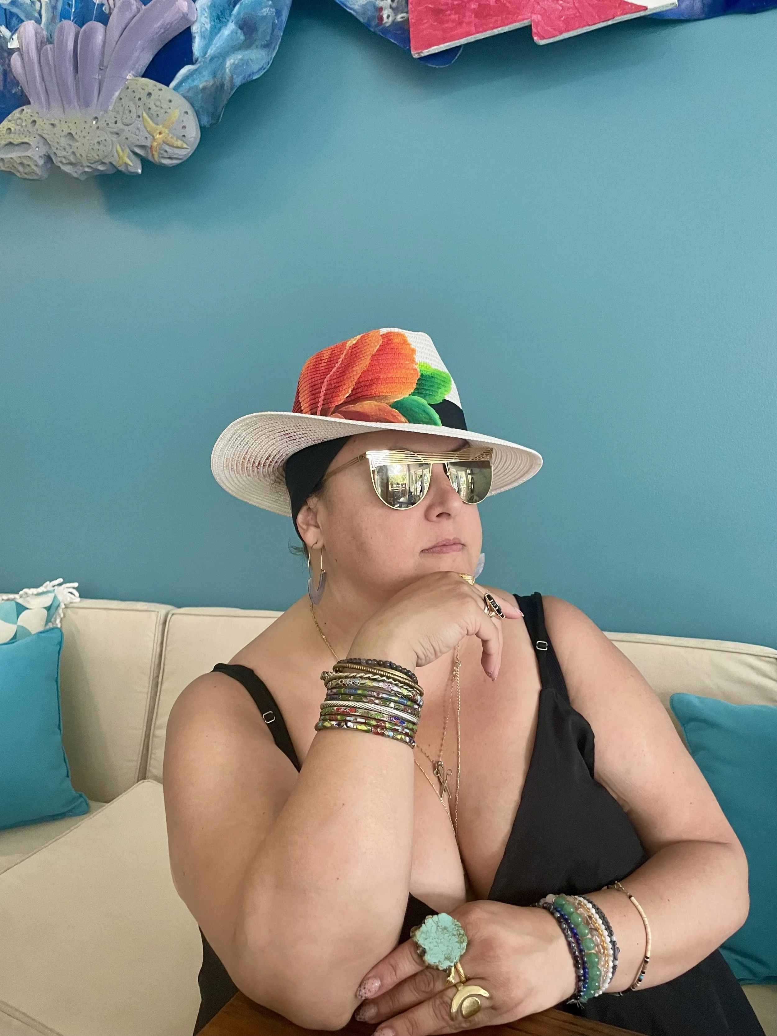 A woman wearing reflective sunglasses, a wide-brimmed hat with a tropical flower pattern, and a black sleeveless top. She is sitting on a cream-colored couch with blue pillows, with her right arm resting on a table, wearing multiple colorful bracelet