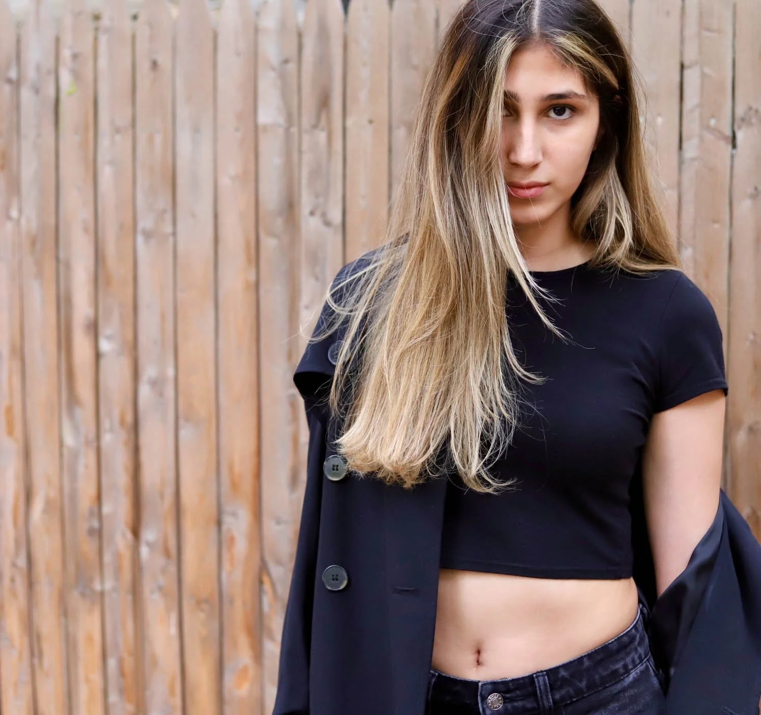 A young woman with long blonde hair wearing a black crop top and black jeans standing in front of a wooden fence.