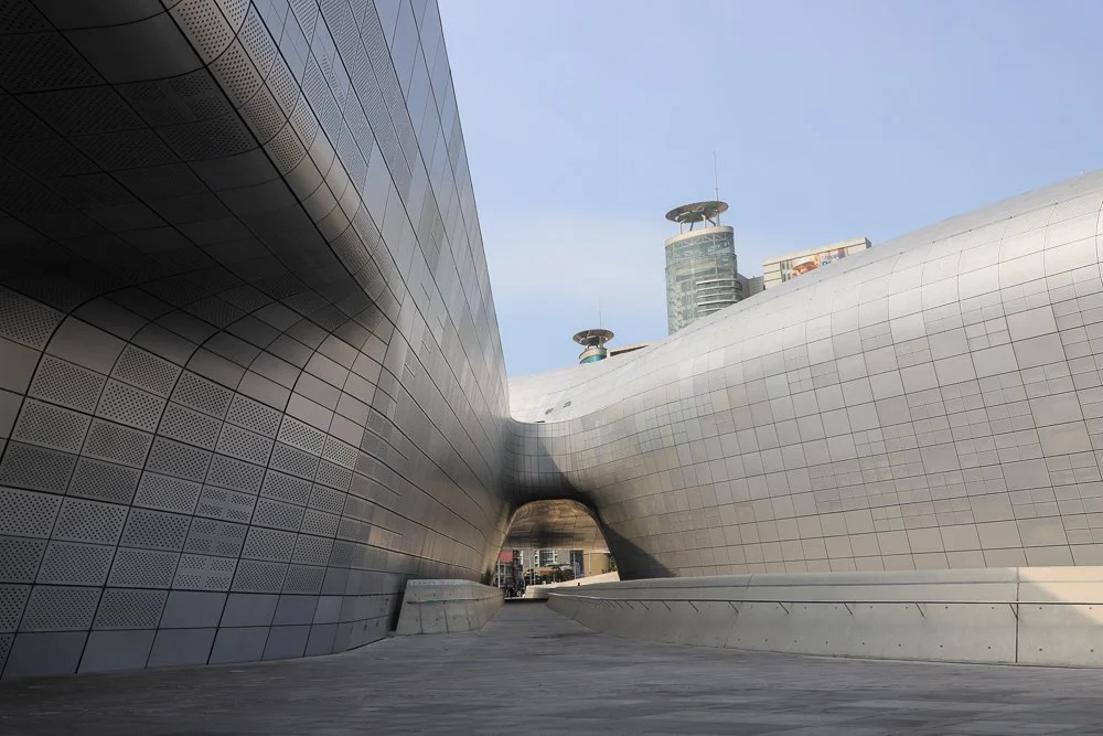 Modern architectural buildings with metallic, curved surfaces and a passageway between them in an urban setting.