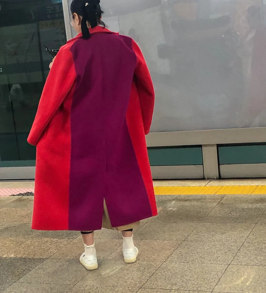 Person wearing a long red and purple coat, beige pants, white shoes, and white socks, standing on a tiled floor in front of a reflective glass wall.