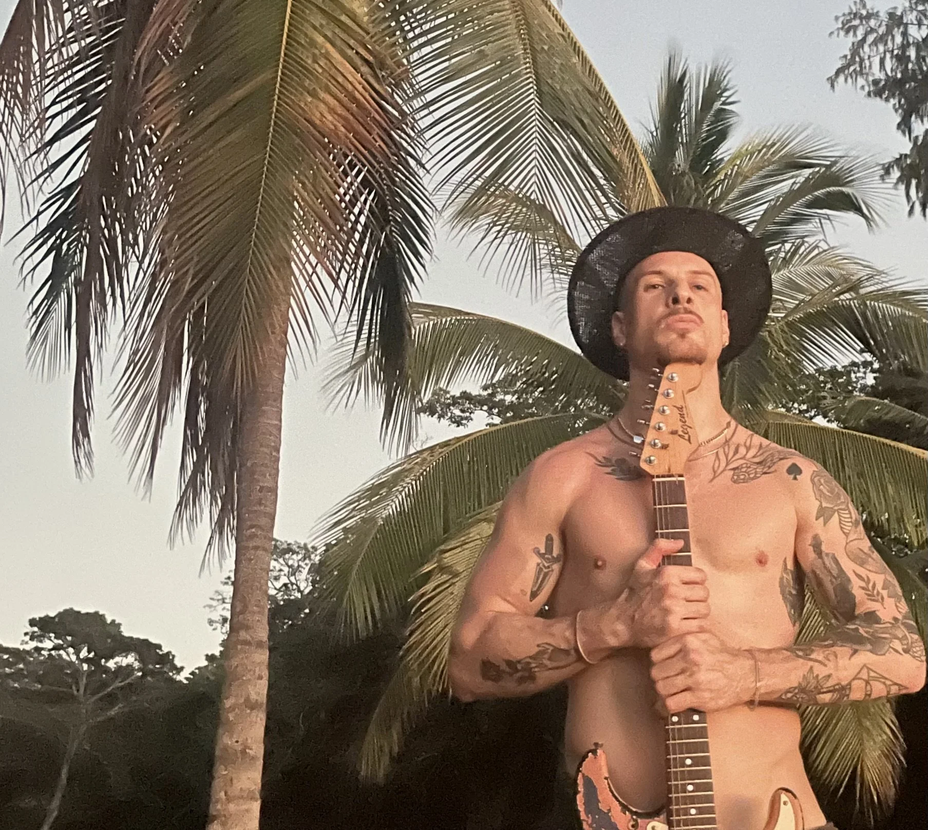 A shirtless man with tattoos holding a guitar, standing outdoors near palm trees at sunset or dusk, wearing a black hat with a wide brim. Roatan.