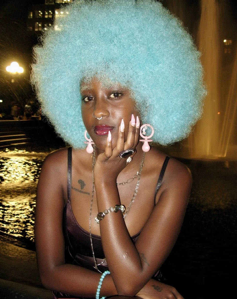 Woman with bright blue afro wig wearing earrings, rings, bracelets, and a satin dress, posing with her hand near her face at night, with city lights and water fountain in background.