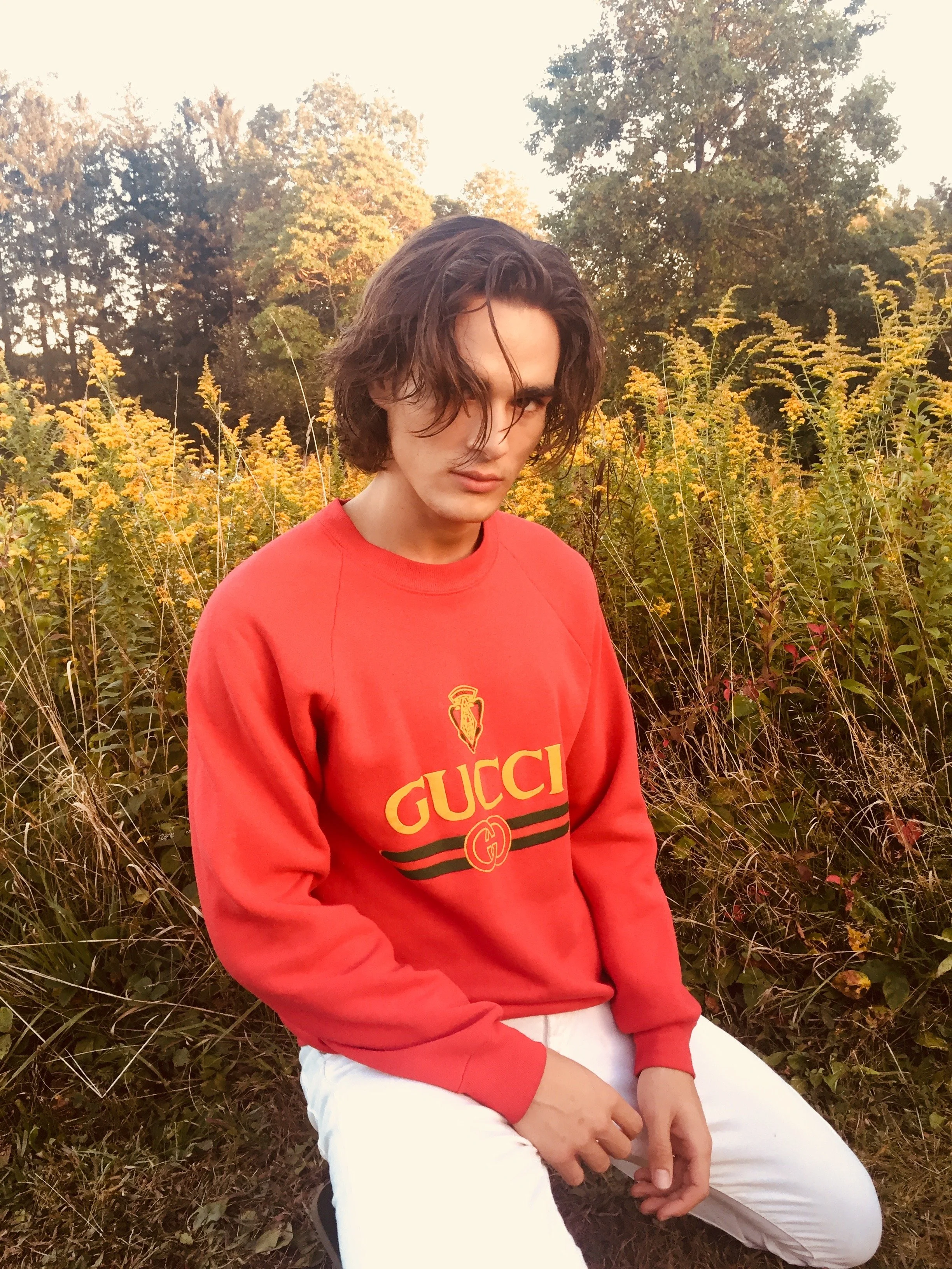 A young man with shoulder-length wavy hair is sitting outdoors in a field with tall yellow flowers and trees in the background, wearing a red Gucci sweatshirt and white pants.