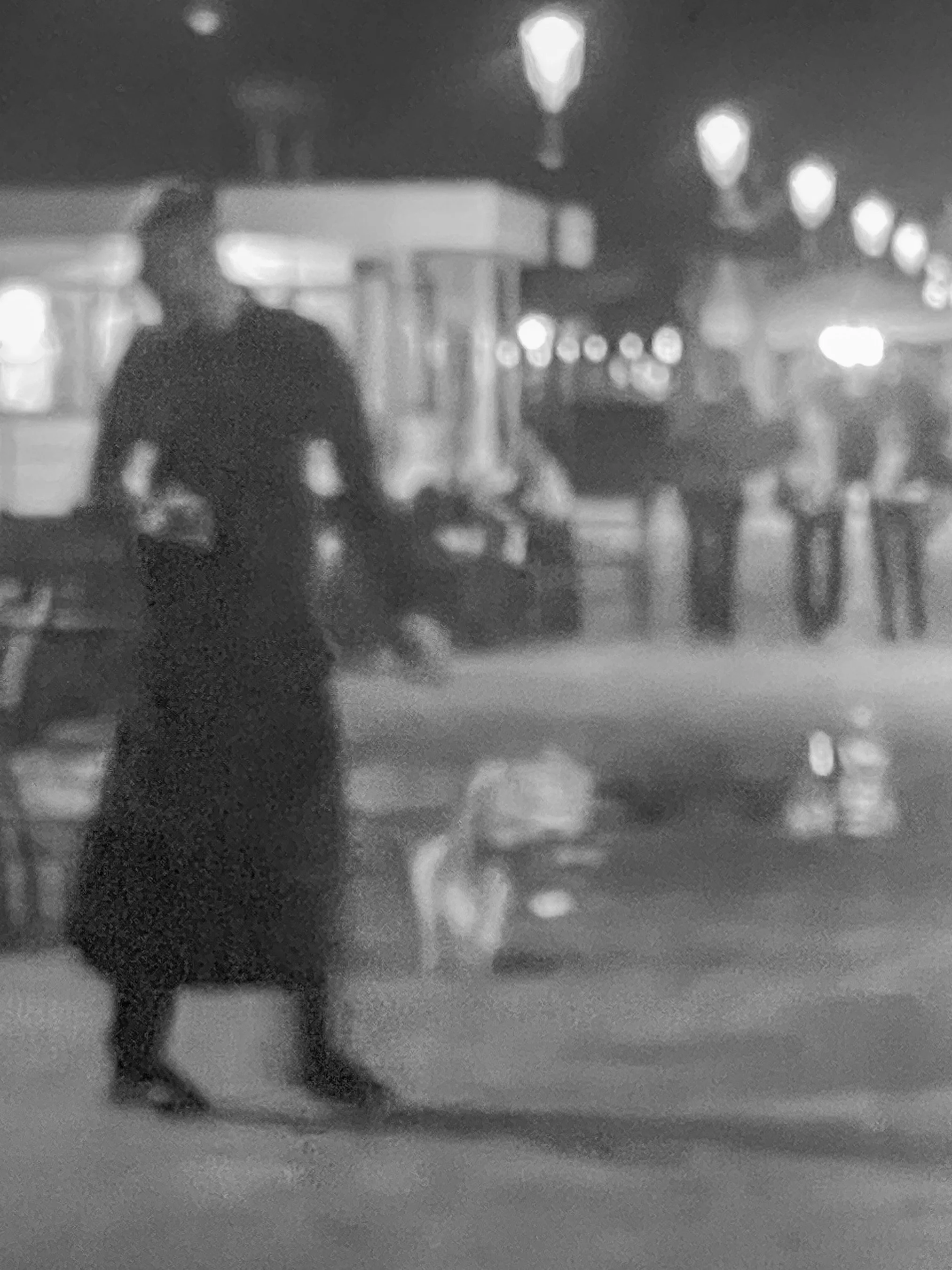 A woman in a dress walking along a street at night with streetlights and blurred figures in the background.