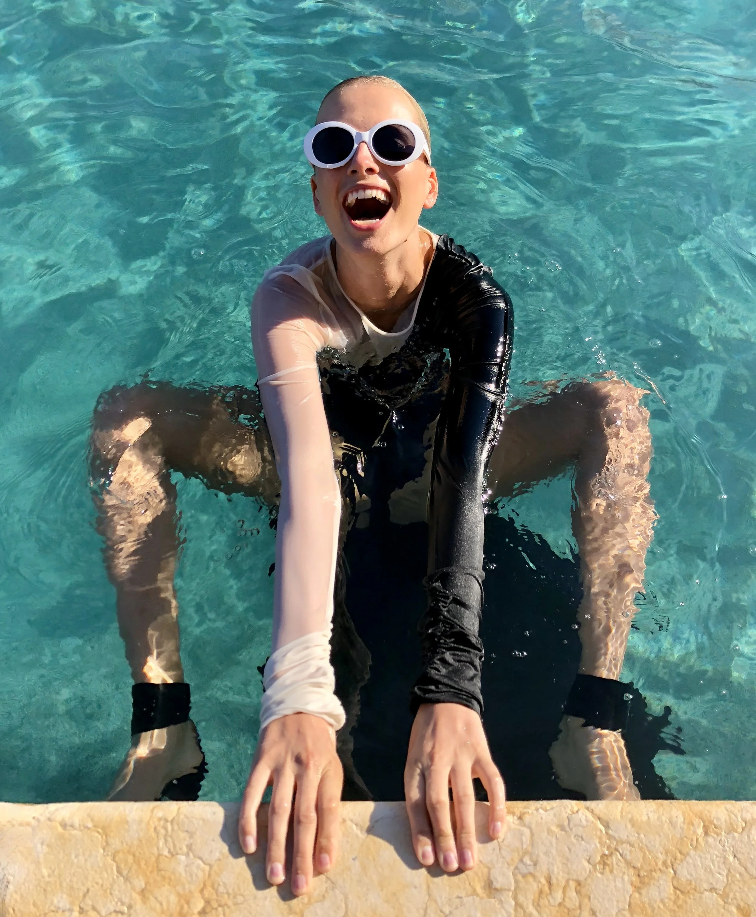 Person in a swimming pool wearing large white sunglasses, a sheer long-sleeve shirt, and black pants, smiling with arms resting on the pool edge.