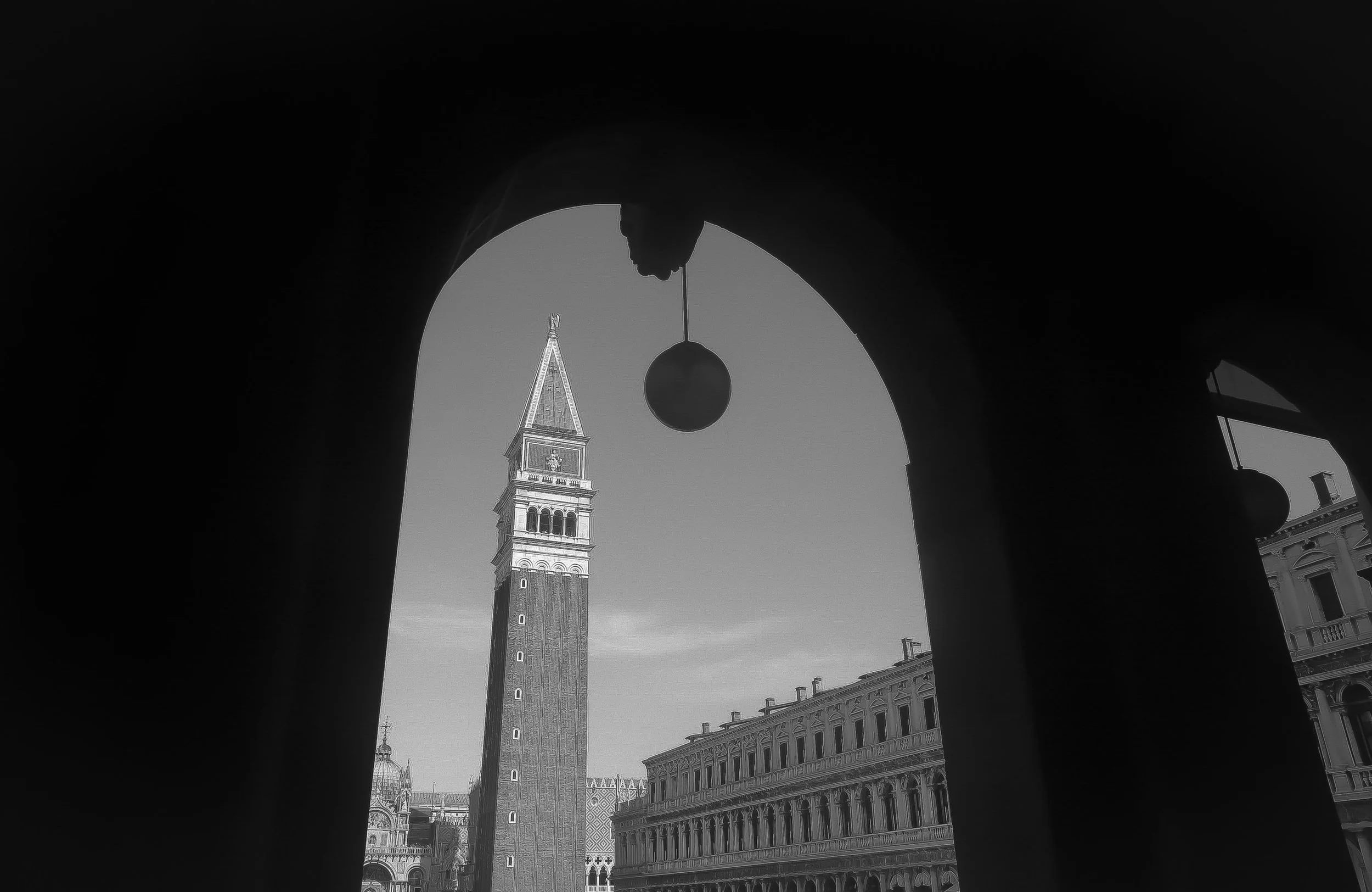 View of Venice's St. Mark's Campanile through an arched structure, with a bell hanging in the foreground and historic buildings in the background.