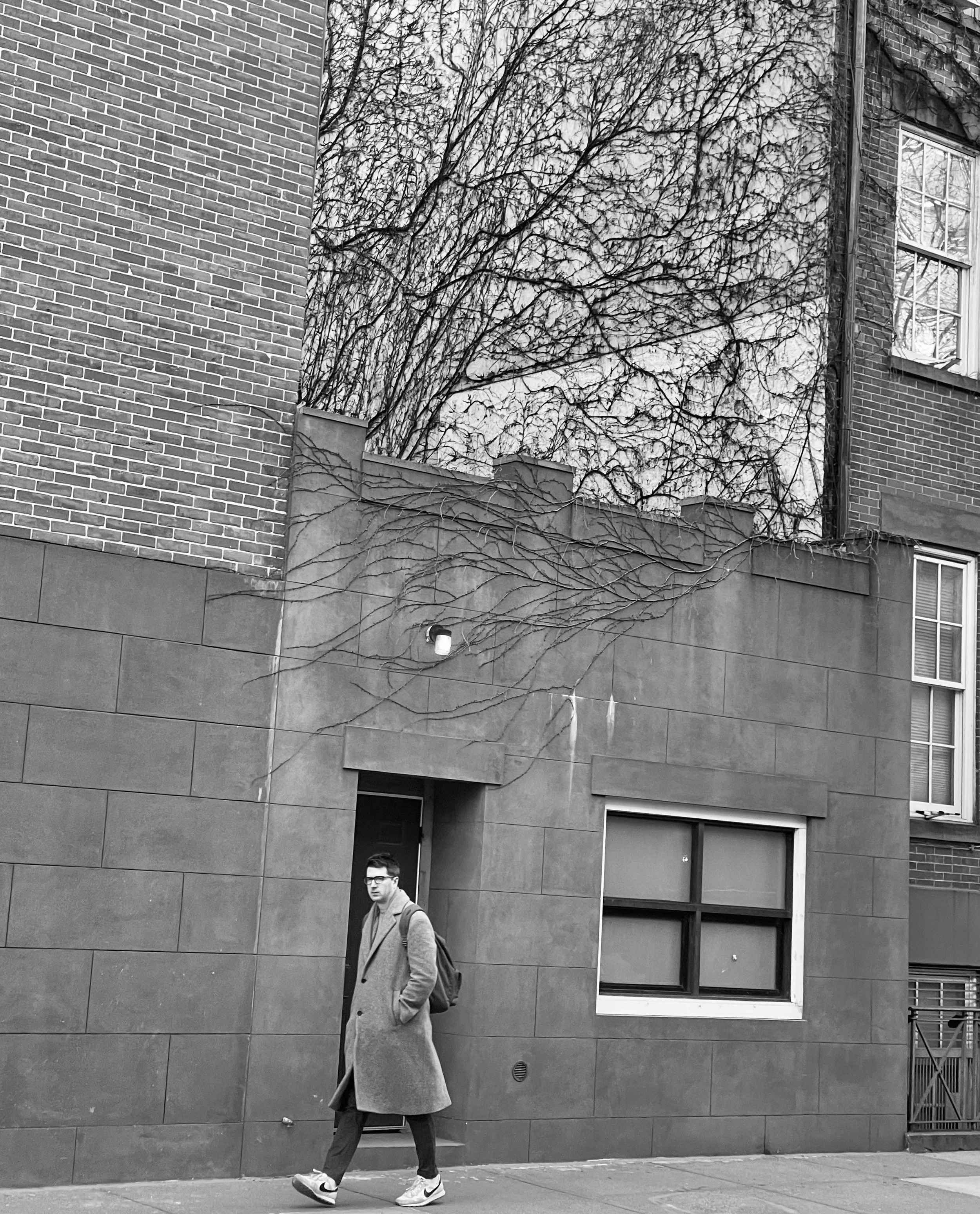 A man in a long coat and sneakers walking on a city sidewalk, with an apartment building and leafless tree branches in the background.