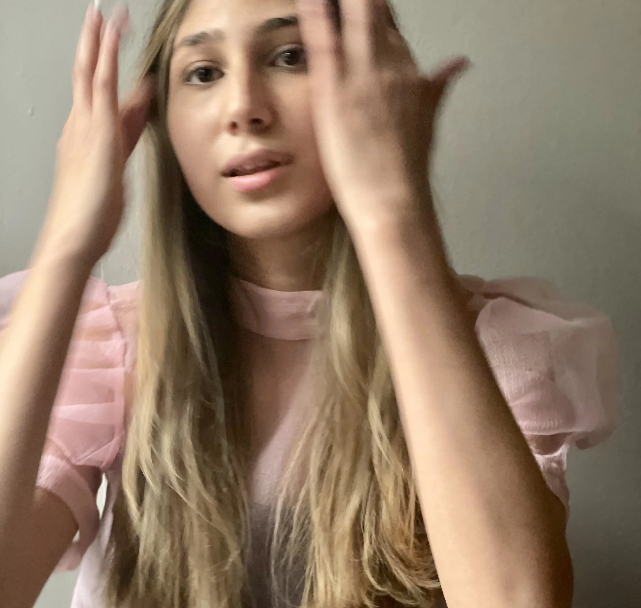 A young woman with long blonde hair wearing a pink sheer top, touching her forehead with both hands, looking into the camera.
