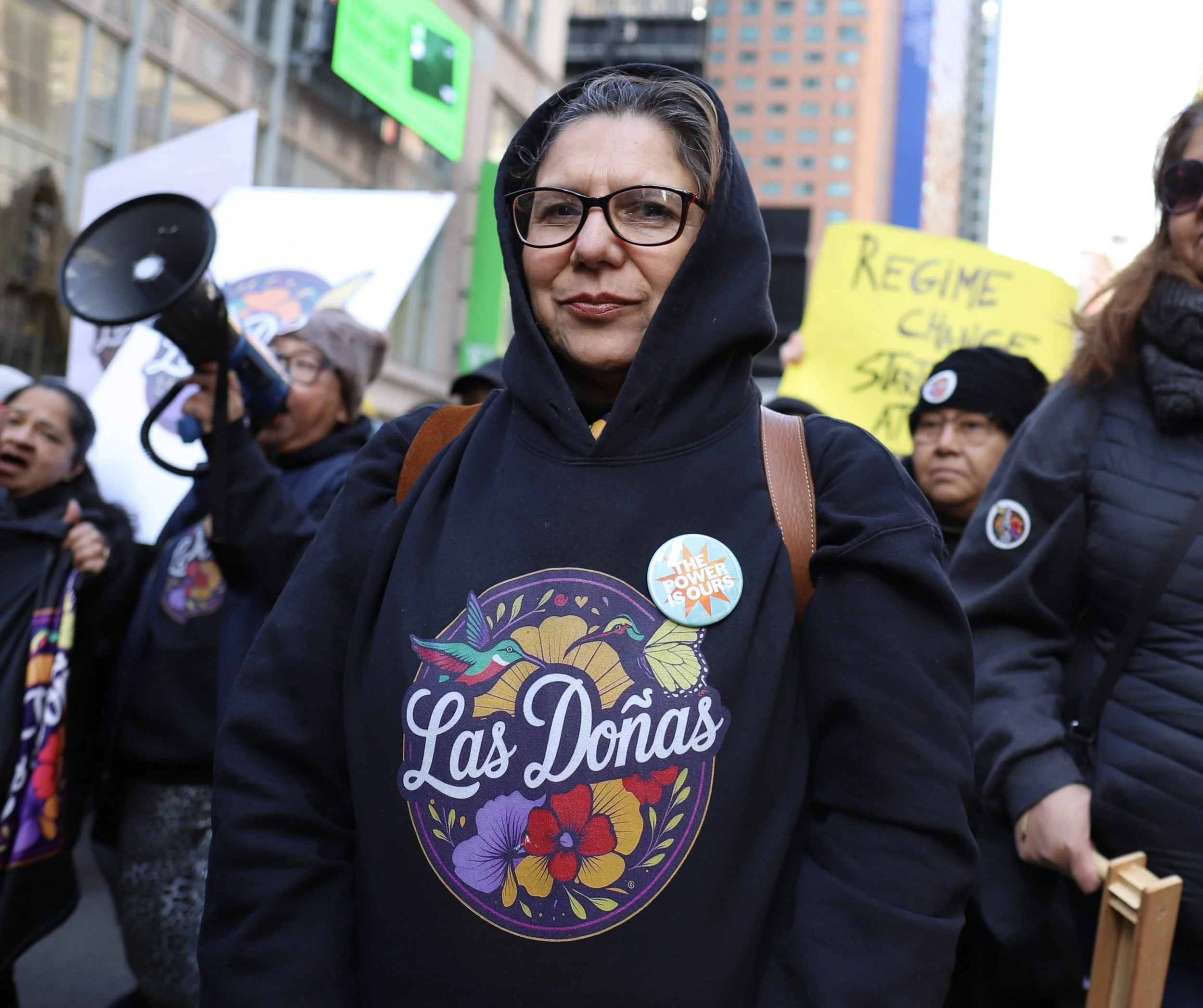 A woman wearing glasses and a black hoodie with a "Las Donas" logo stands at a protest march. She has a round badge saying "The Power is Ours." Around her, protesters hold signs and a megaphone, with city buildings in the background.