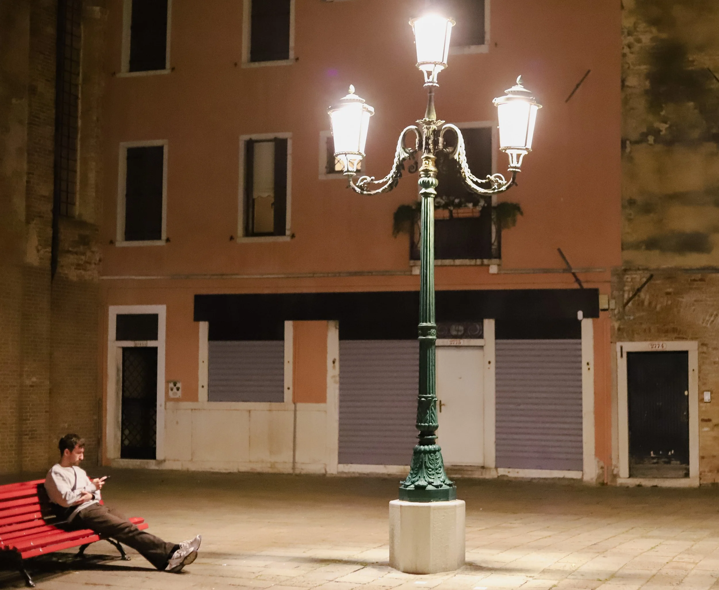 A young man is sitting on a red bench at night, looking at his phone, with a large, ornate streetlamp illuminating an empty city square with closed shop shutters and residential buildings in the background.