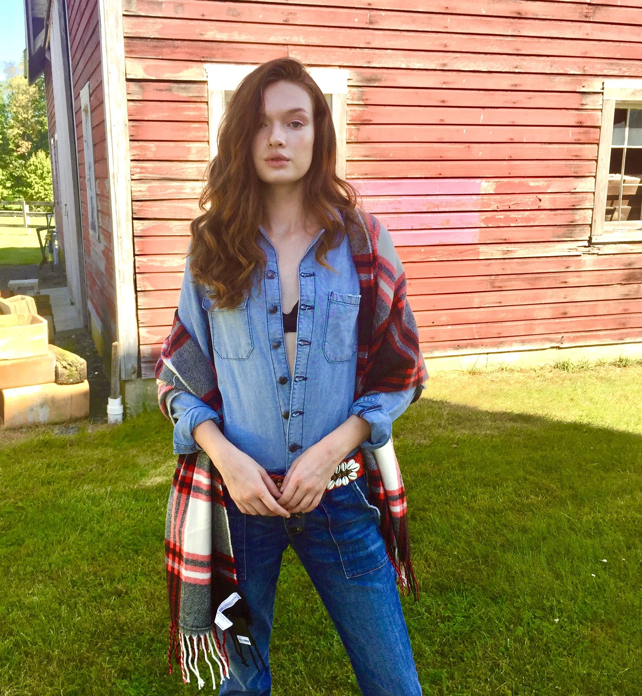 A young woman with long, wavy auburn hair standing in front of a red wooden building with a window. She is wearing a denim shirt layered over a black top, with a red and black plaid scarf draped over her shoulders, and blue jeans. She has her hands c