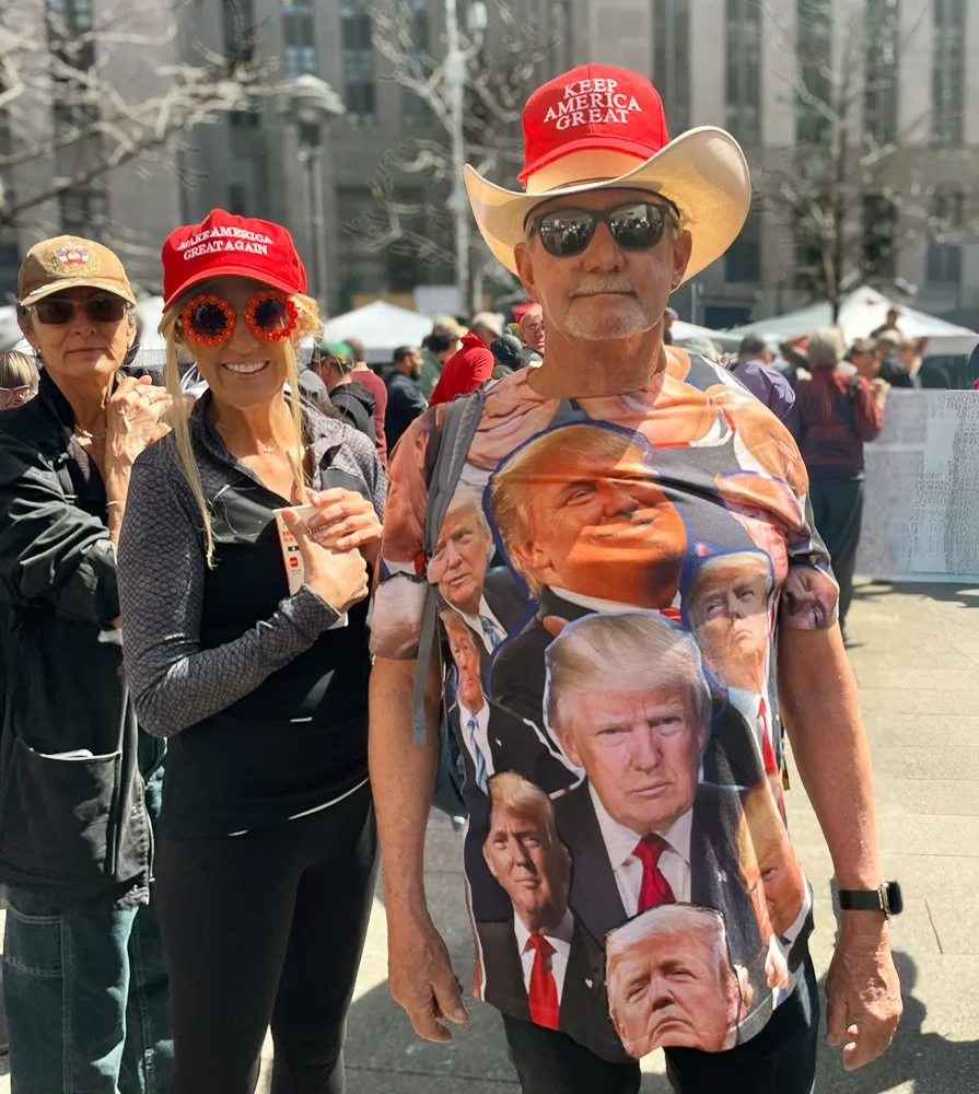 A man wearing a Donald Trump shirt and a red hat with 'Keep America Great' at a rally or protest, with two women in the background also wearing red caps and one of them wearing festive sunglasses, in an outdoor urban setting with other people and ten