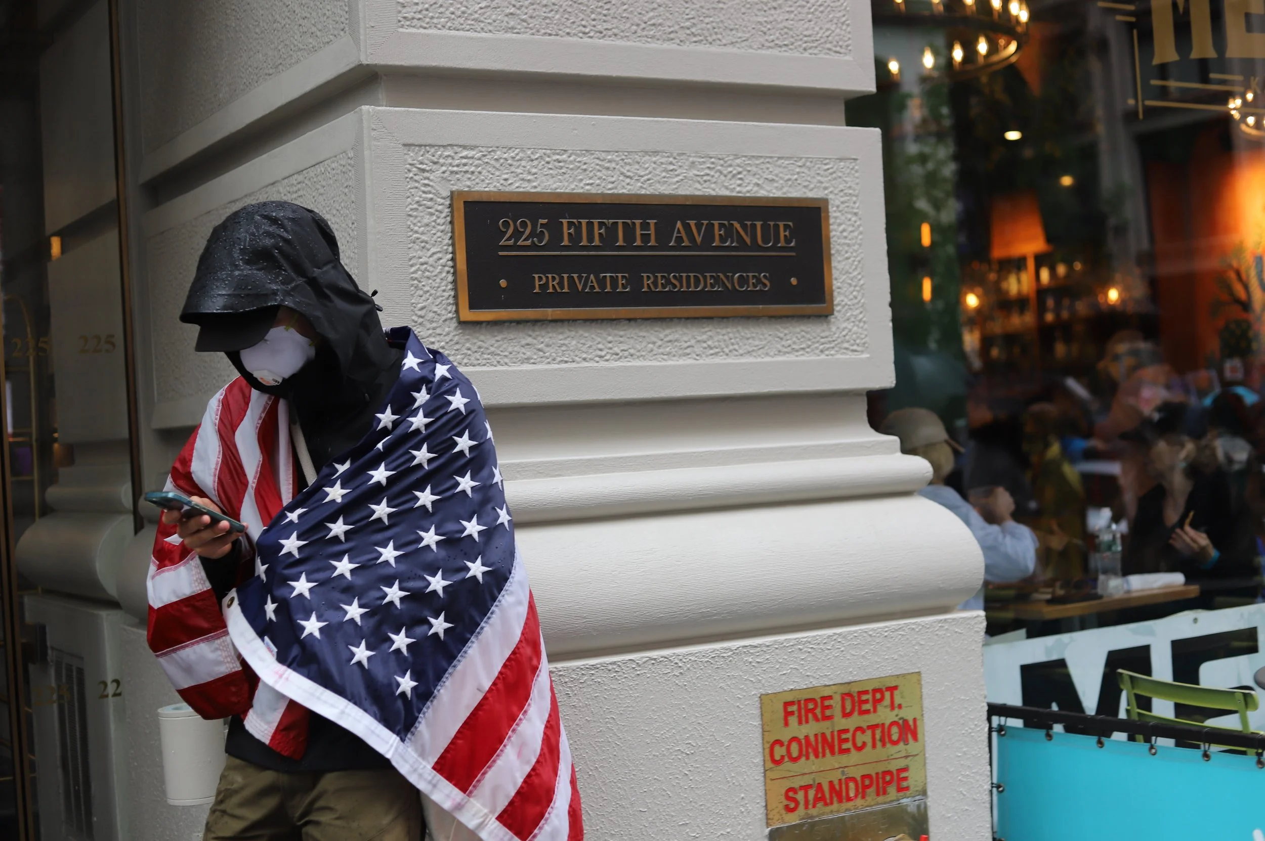 Person wearing a face mask with water droplets, wrapped in an American flag, standing next to a building with a sign that reads '225 Fifth Avenue, Private Residences.' The person is looking at a smartphone. There is a reflection of people dining insi