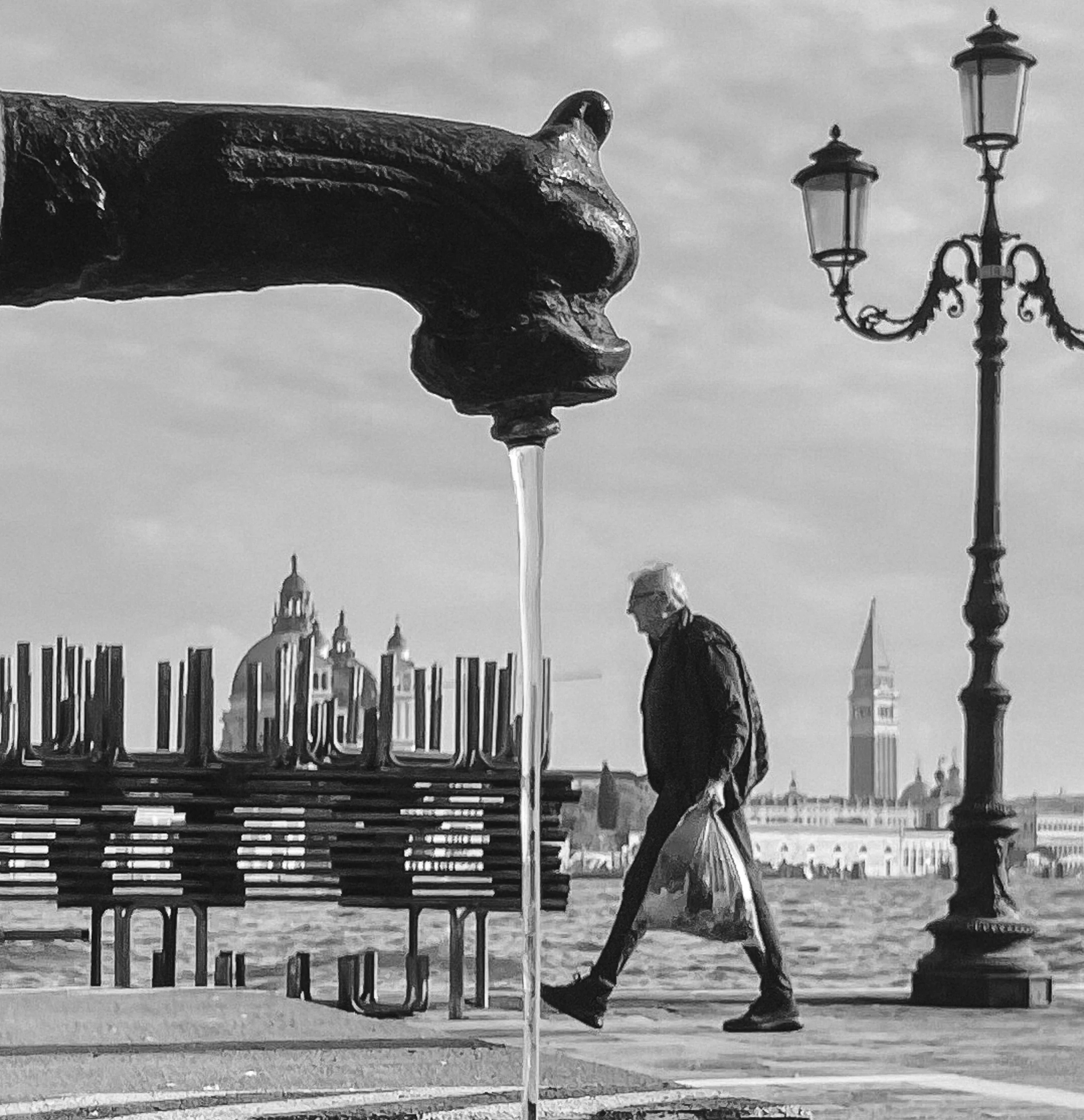 A black and white photo of a street scene in Venice, Italy. The foreground shows a close-up of a street fountain with water flowing from a spout. In the background, an elderly man walks past, carrying a plastic bag, with the iconic St. Mark's Campani