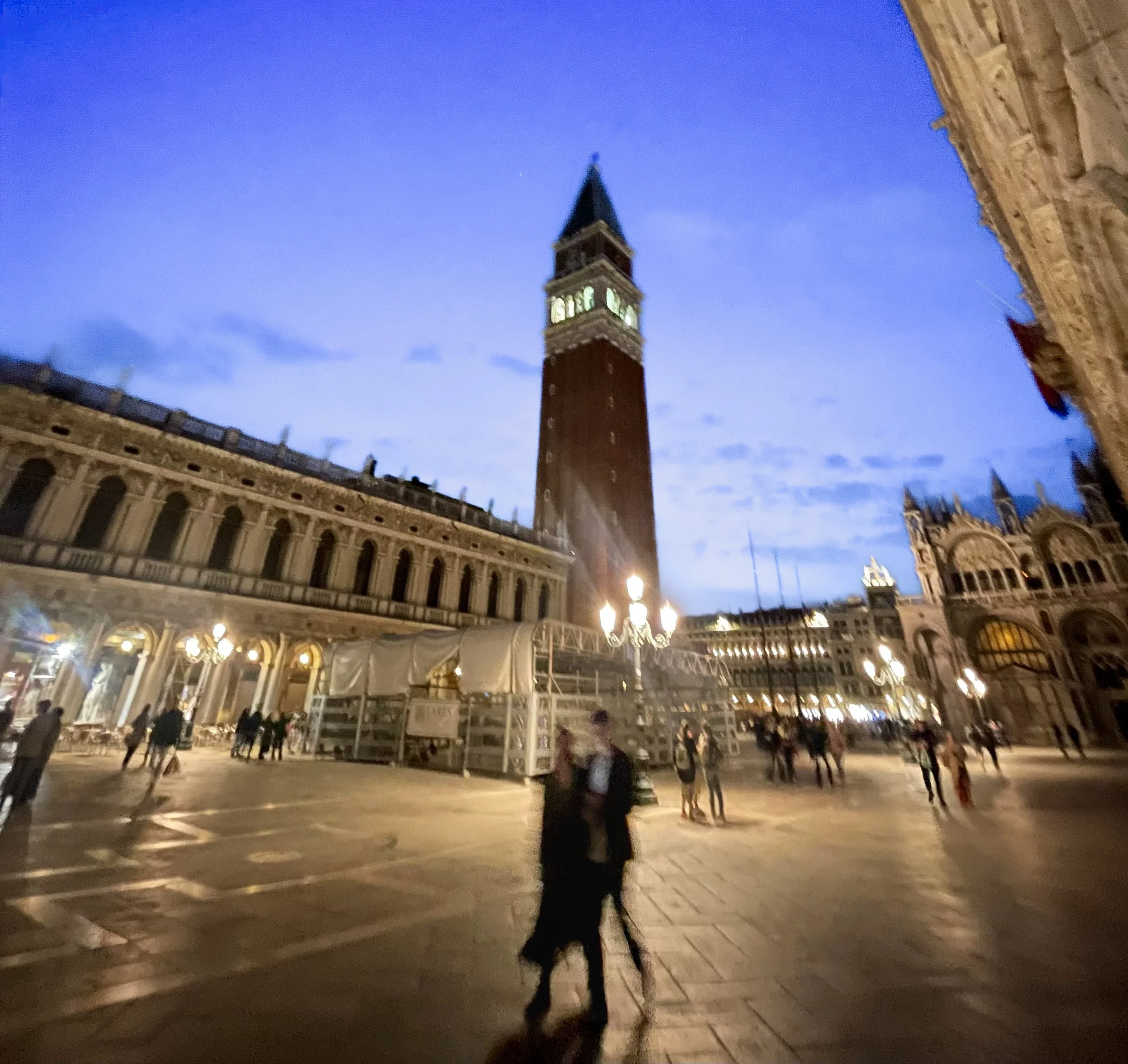 Nighttime view of Piazza San Marco in Venice, Italy, with the Campanile di San Marco tower illuminated and people walking in the square.