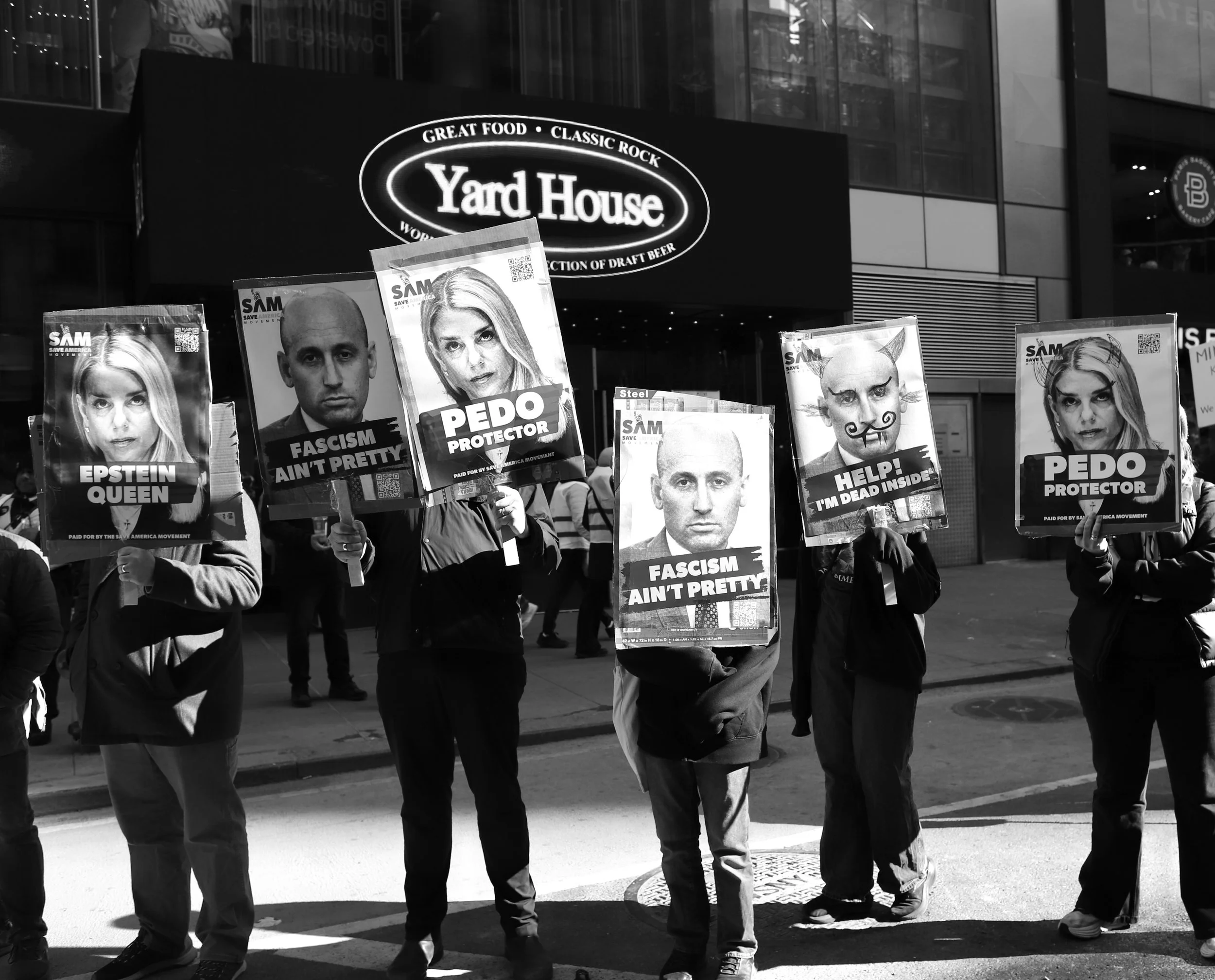 People holding protest signs with images and messages like 'Epstein Queen', 'Fascism Ain't Pretty', 'Pedophile Protector', and 'Help! I'm Dead Inside' in front of a city building with a large sign for Yard House.