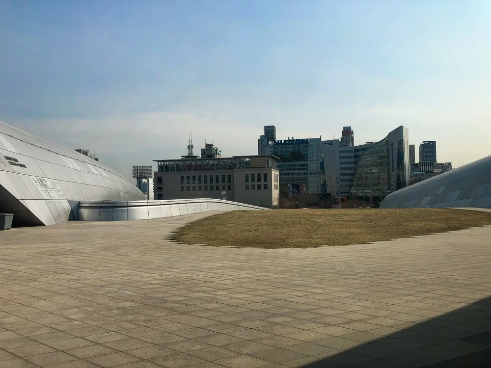 Modern cityscape with distinctive curved metallic buildings and a grass-covered area in the foreground.