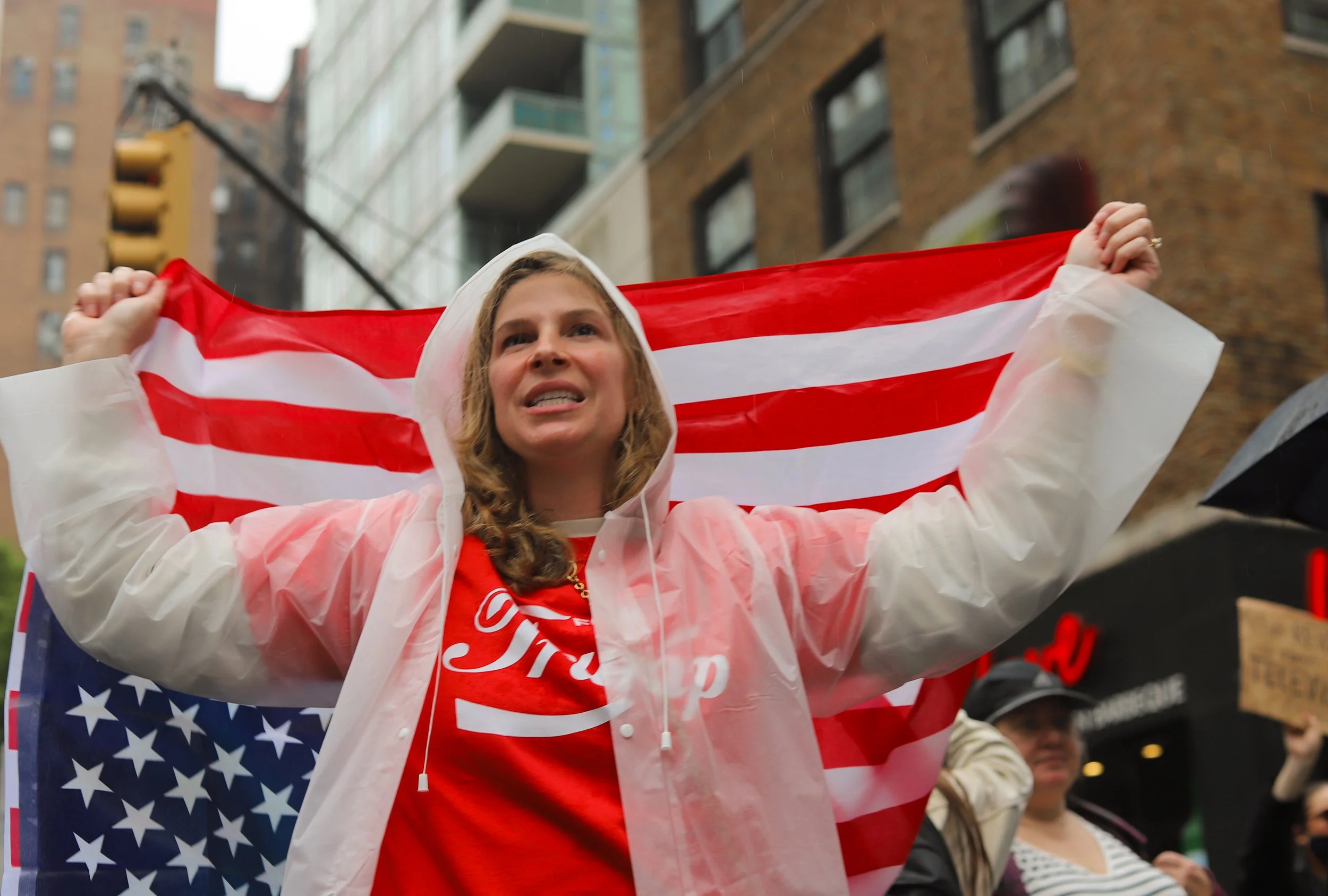 Woman holding an American flag at a protest or rally, with buildings and other protesters in the background.