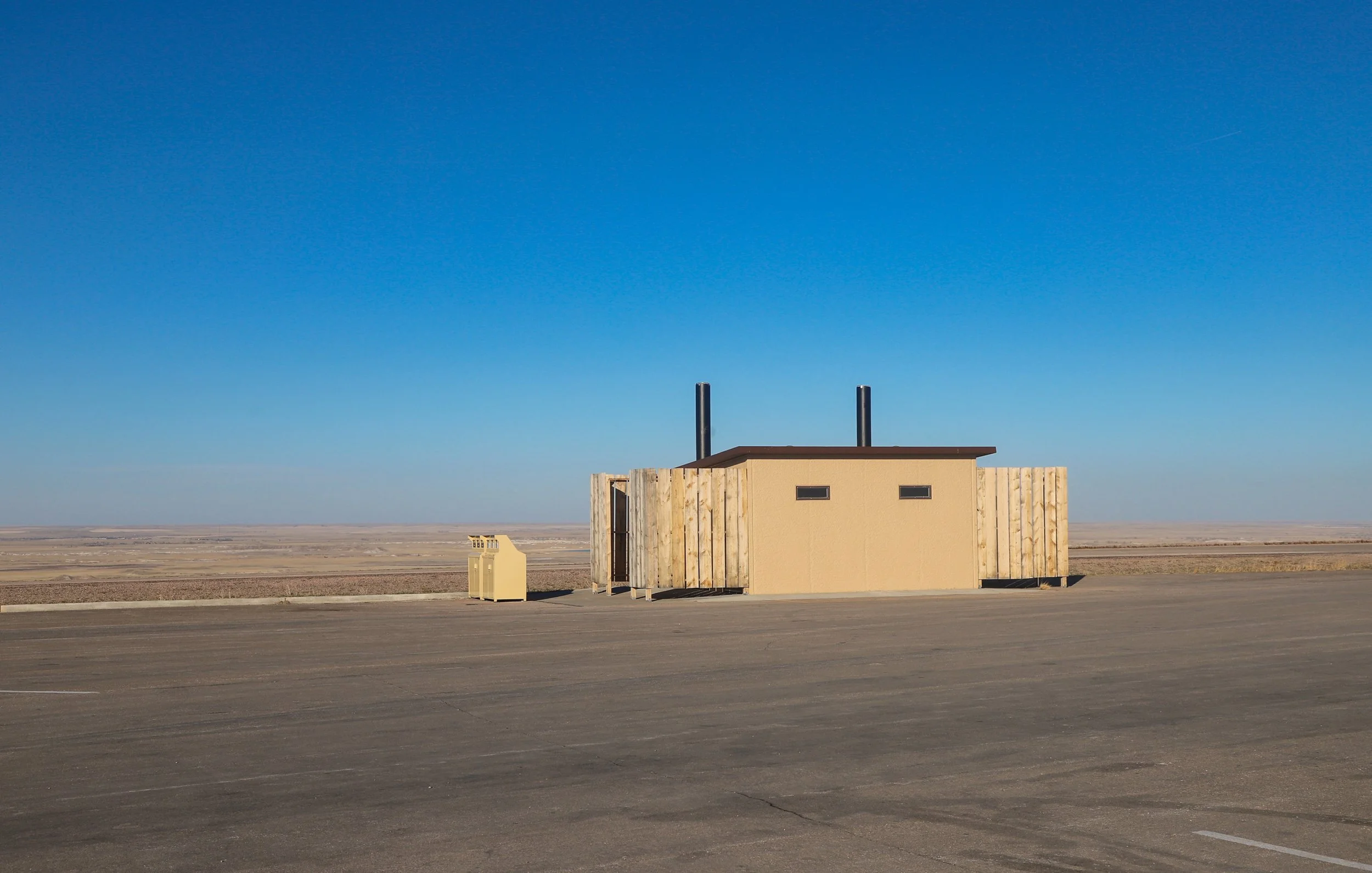 A small beige building with black vents and two black chimneys in a large, empty parking lot in a flat, desert landscape under a clear blue sky.